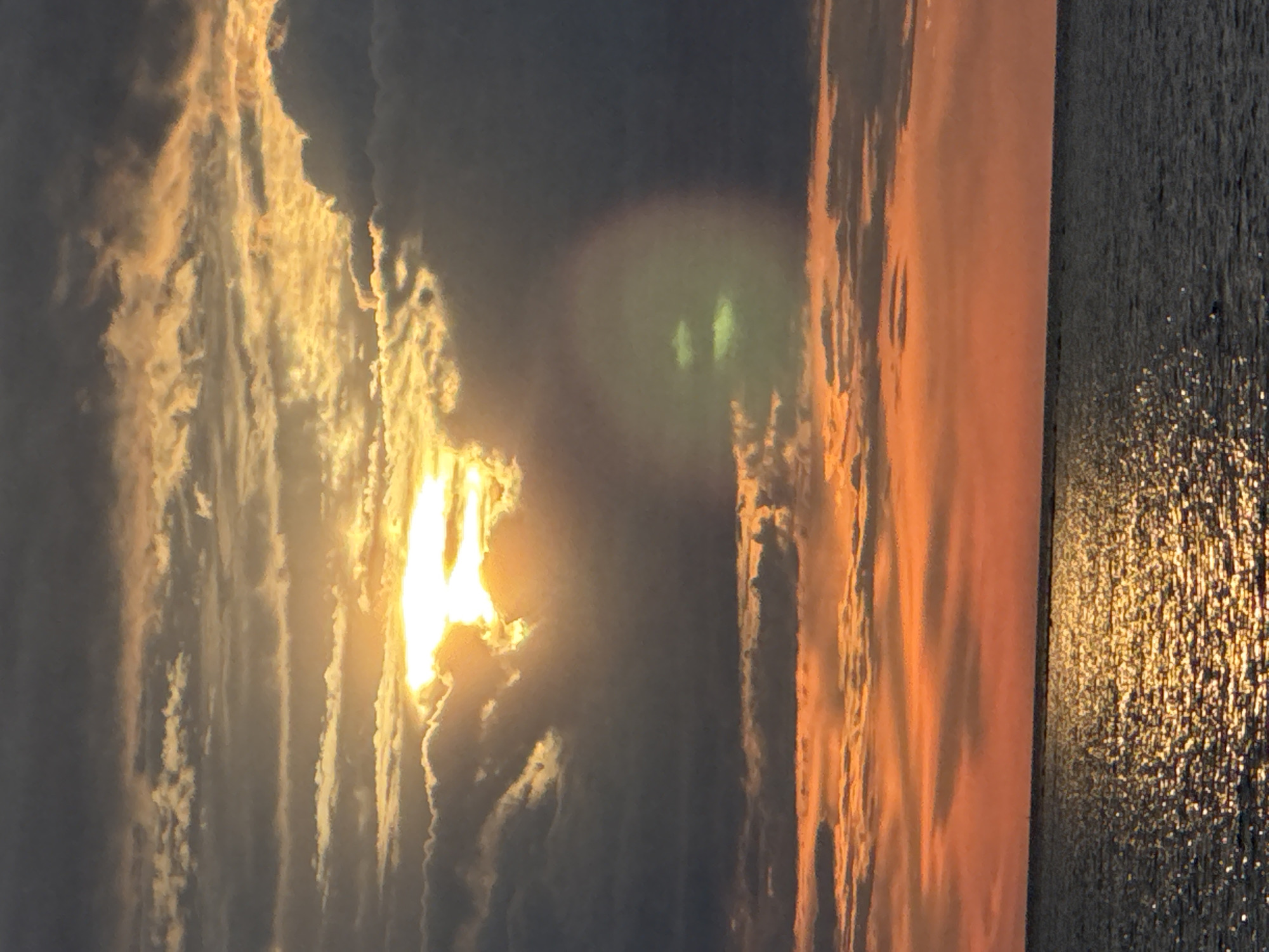 This image depicts a sunset over the Baltic Sea, with the silhouette of Tallinn, Estonia, visible in the distance. The sky is a dramatic display of color, with hues of orange, yellow, and grey blending together as the sun dips below the horizon. Wispy clouds are illuminated by the setting sun, creating a beautiful contrast between light and shadow. The sea reflects the colors of the sky, creating a shimmering, golden path that leads towards the distant city. The scene is tranquil and serene, evoking a sense of peace and natural beauty.