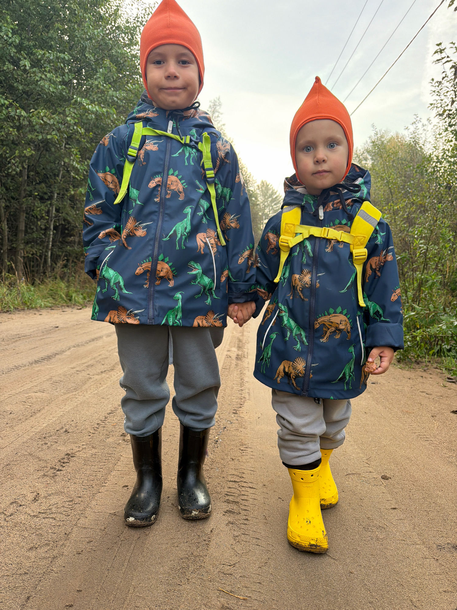 Two young children, a boy and a younger boy, stand on a dirt road in a wooded area. The older boy, on the left, is wearing an orange beanie, a dark blue jacket with a dinosaur print and bright green accents, gray sweatpants, and black rubber boots. The younger boy, on the right, wears a matching orange beanie, a similar dinosaur-print jacket with yellow straps, gray sweatpants, and yellow rubber boots. They are holding hands. The younger boy is picking up a small stone. The road appears to be a rural track, possibly in or near the city of Chaikovsky, Russia, indicated by the surrounding trees and the overcast sky. The overall scene suggests an outdoor excursion or walk in nature.