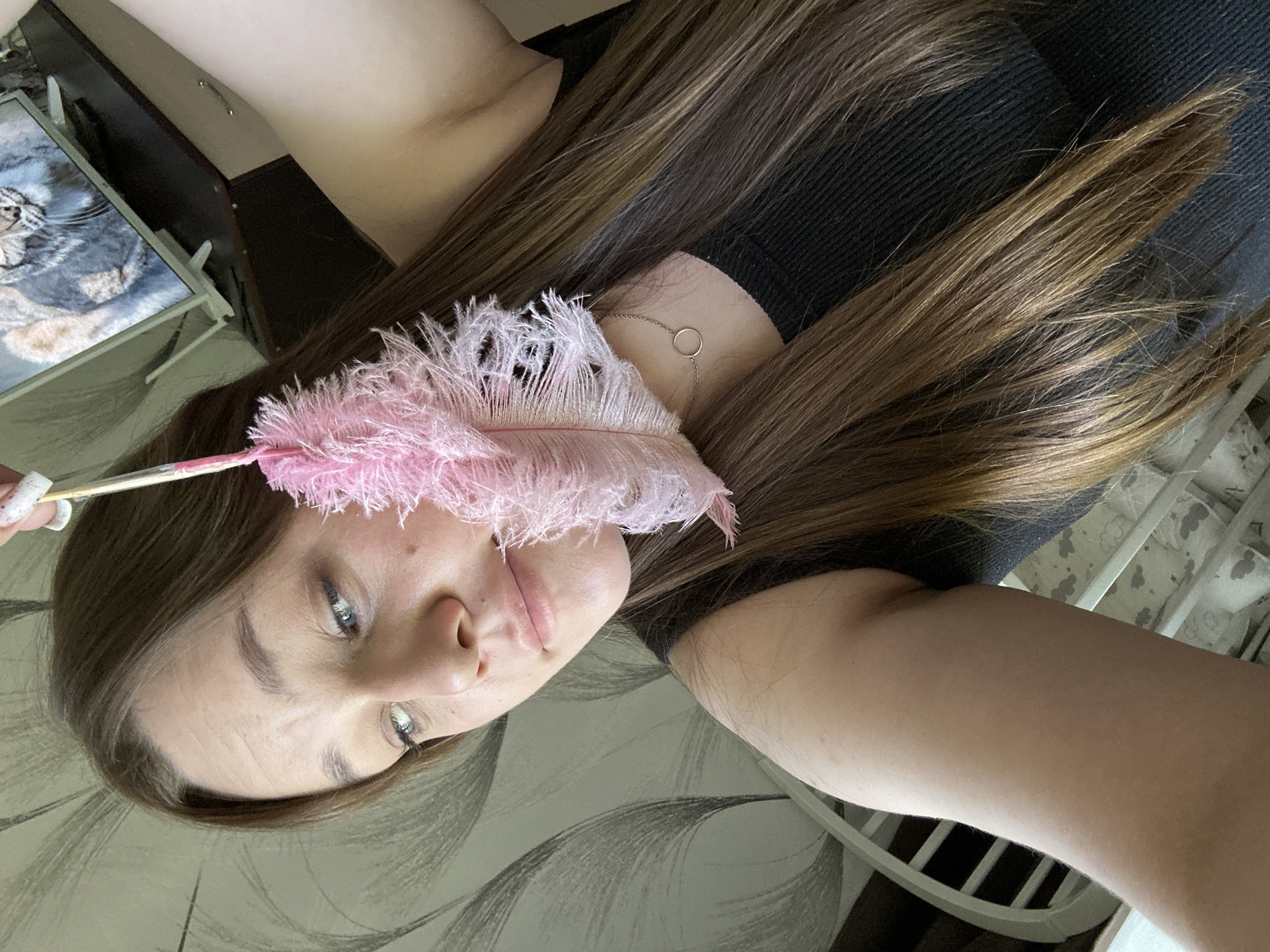 A person with long, light brown hair and fair skin is holding a pink feather duster near their face. The person is wearing a black top and a delicate silver necklace. The background includes a patterned wall and a glimpse of a picture frame with an image of a cat. A white crib with a patterned interior is also visible in the background. The location is Chaikovsky, Russia.