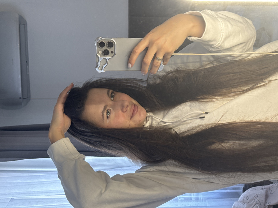 The image depicts a mirror selfie of a lone female individual in an indoor room, located in Molodi, Russia, as specified. The subject, a woman with long, dark brown hair, is positioned in the center of the frame, wearing a white hooded sweatshirt with visible black drawstrings. Her left hand is raised, fingers splayed and resting on the top of her head, appearing to adjust her hair. Her right hand holds a silver-colored smartphone, which is encased in a translucent protective case featuring a distinct irregular silver-toned frame around the camera array. The phone shows three visible camera lenses on its rear and has a white charging cable connected and hanging downwards. A plain silver-colored ring is visible on the ring finger of her right hand. Her fingernails are manicured with a light polish. She exhibits a subtle smile, looking directly into the smartphone's lens, which is reflecting her image.

The background, visible in the mirror's reflection, includes a light-colored curtain or sheer fabric on the upper left. A silver-grey wall-mounted air conditioning unit is positioned on the upper right. A vertical strip of bright artificial light is present along the right edge of the frame. On the lower left, a crumpled white fabric with faint, light-colored patterns is partially visible. The overall environment suggests a private room, possibly a bedroom or living space, with light-toned walls.