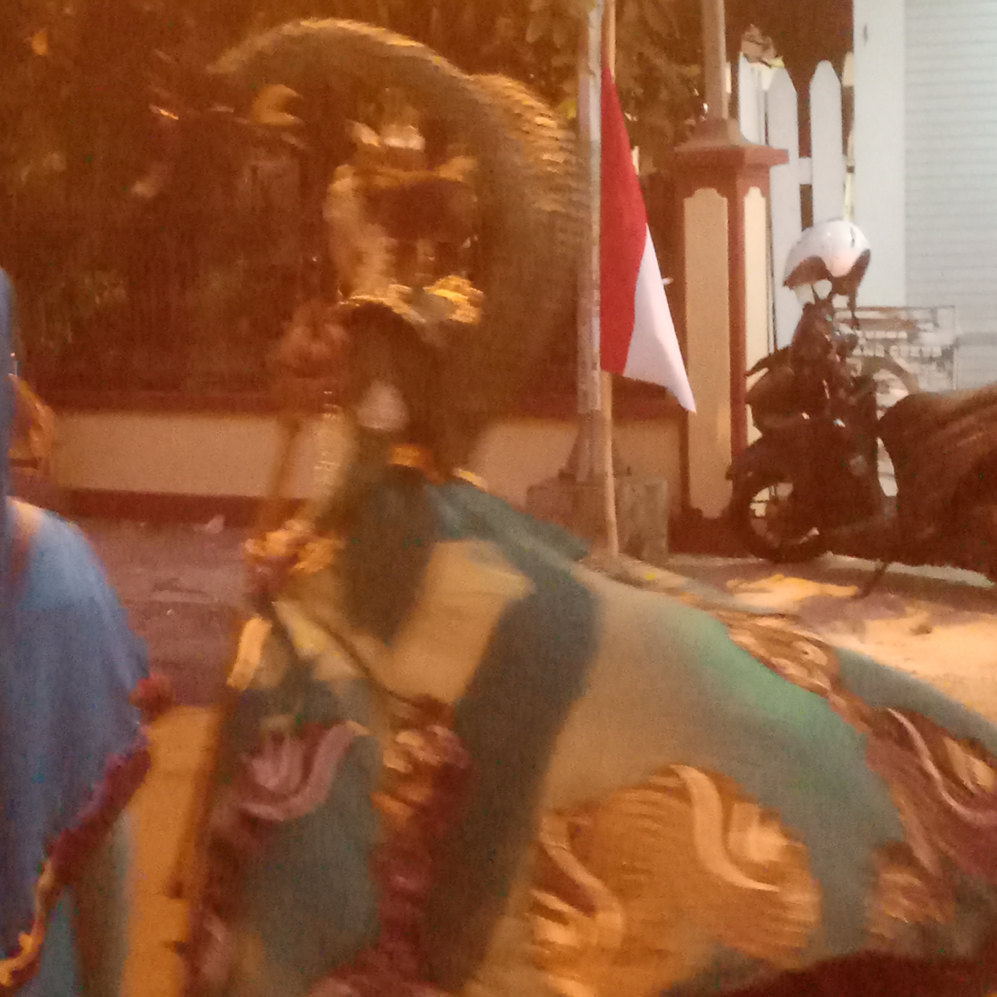 The image depicts a street scene in Semanding, Indonesia, possibly at dusk or night given the lighting. In the foreground, a large, multi-colored fabric, possibly part of a costume or decoration, is draped and angled across the frame. The fabric has patterns in shades of green, beige, and brown. To the left, a person wearing a blue garment with reddish trim is partially visible. In the mid-ground, a motorcycle with a white helmet resting on its seat is parked. Behind the motorcycle, a white picket fence and a building are visible. A pole with the Indonesian flag, red and white, is planted near the motorcycle, suggesting a national celebration or event. The background is indistinct, appearing to be foliage or trees, with warm lighting illuminating the scene. The overall impression is of a community gathering or event taking place on a street.