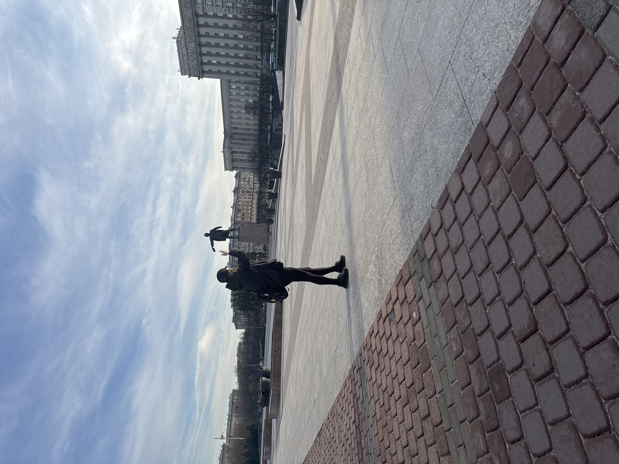 An eye-level photograph, oriented sideways, shows a figure standing near a statue in an urban setting. The figure, dressed in a dark-colored outfit, stands with one arm raised, appearing to interact with the statue above, which is also dressed in a similar color. A backpack is visible on the figure's back.
The ground is composed of gray pavers and reddish-brown brick pavers. Behind the figure, there are several buildings. A large gray building dominates the background, with smaller structures alongside it.
The sky is visible, with wispy clouds.
Based on the style of architecture, particularly the large gray building, the location is likely in Saint Petersburg, Russia, near the Moskovsky Victory Park and the House of Soviets. The statue is likely a monument to Lenin.