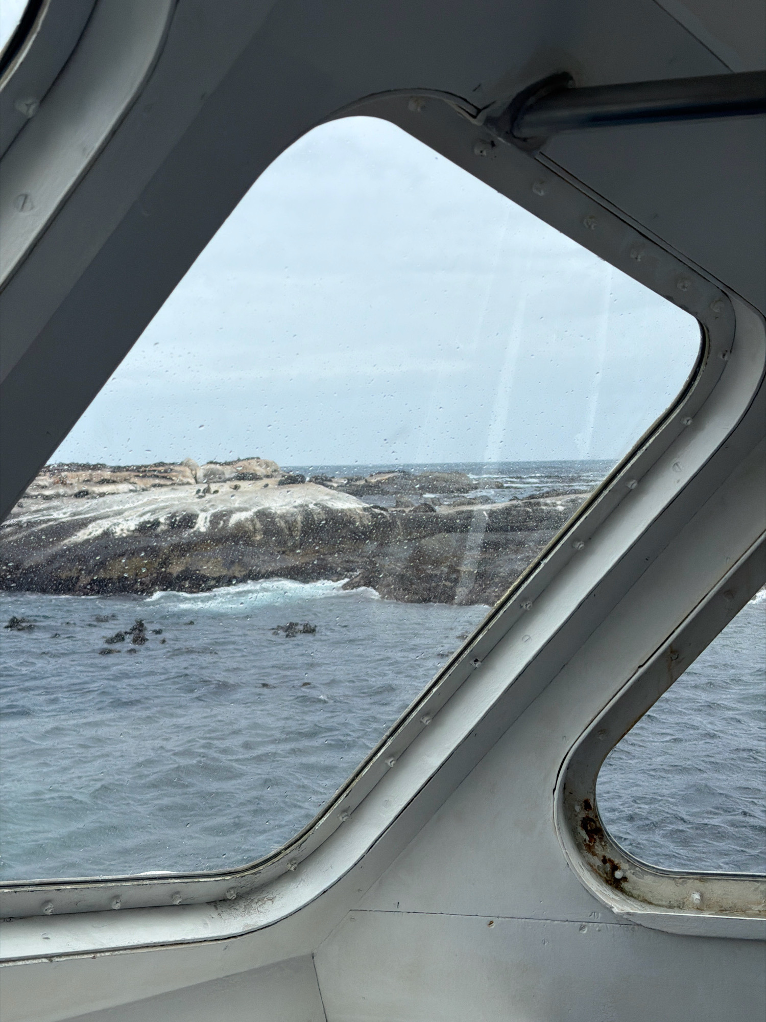 The image captures a view through a boat's window, looking out at a rocky outcrop in the ocean. The window frame is painted white and shows signs of wear and tear, with some rust visible. The glass of the window is speckled with raindrops, indicating a wet or misty day.
Beyond the window, the rocky outcrop is a prominent feature. It is partially covered in what appears to be guano, creating large patches of white against the dark rock. Several dark shapes, consistent with the appearance of seals or penguins, are scattered across the surface of the rocks. The sea around the outcrop is a deep blue-grey, with white foam where waves are breaking against the rocks. In the distance, the horizon is visible, meeting a pale, overcast sky.
This scene is likely located at Duiker Island (also known as Seal Island) off the coast of Hout Bay, South Africa. Duiker Island is a known habitat for Cape fur seals and African penguins, and the rocky, guano-covered appearance of the island is characteristic of such colonies. The view from a boat is also typical of tours that visit the island to observe the wildlife. The overcast and rainy weather is also common in the Cape Peninsula.