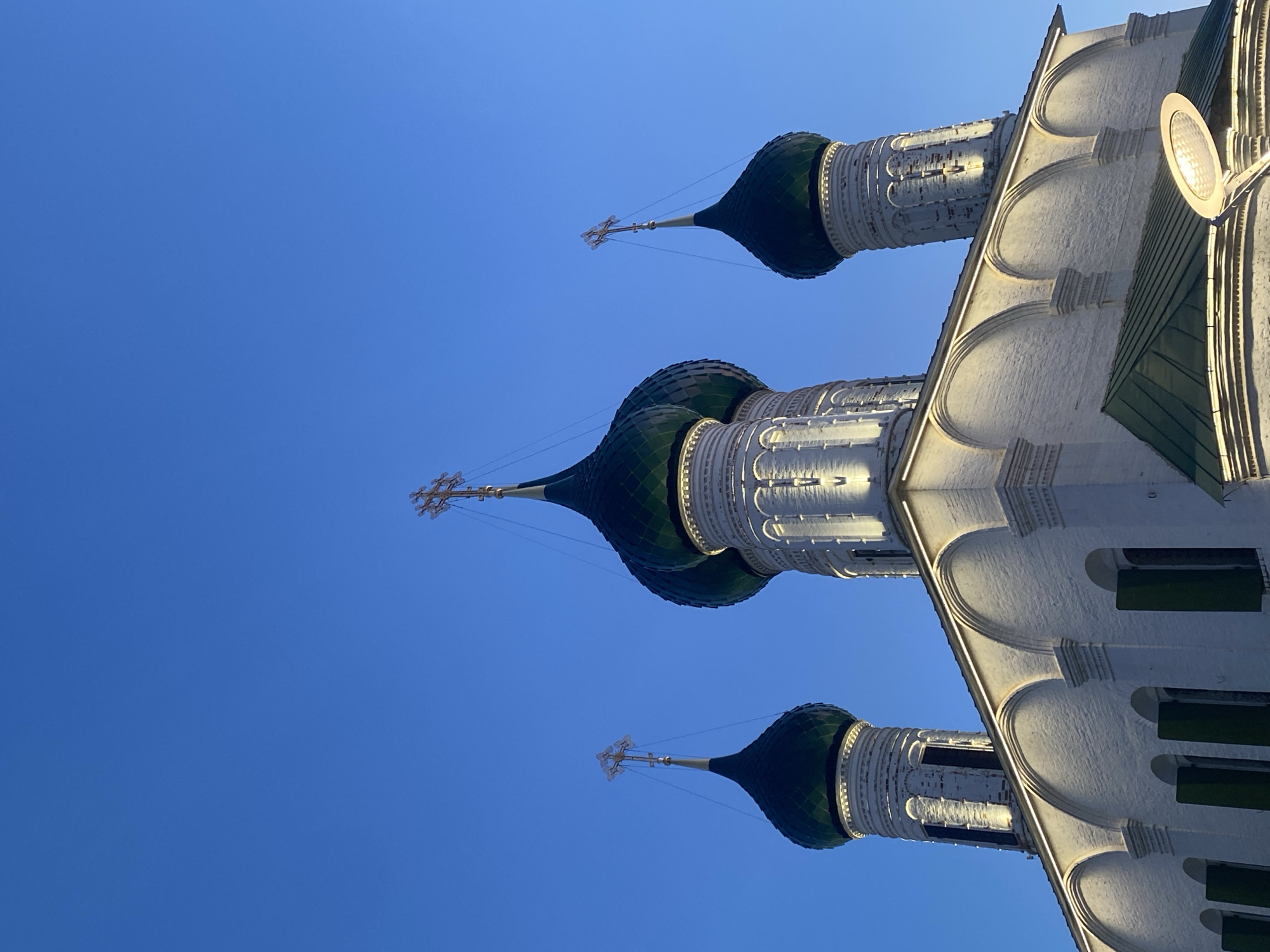 The image displays a low-angle, upward-looking view of the upper part of a large, white architectural structure, likely a church or cathedral, set against a clear, deep blue sky. Three prominent onion-shaped domes, each colored dark green and topped with an ornate gold-colored Orthodox cross, crown the building.
The building's facade, constructed from a light-colored material, features decorative architectural elements, including a series of arched recesses along its upper edge, which cast subtle shadows. The lighting suggests either early morning or late afternoon, with warm sunlight illuminating parts of the structure from the left, creating strong highlights and contrasting shadows that emphasize its texture. A modern, round light fixture with a reflective surface is visible on the upper right side of the building, near one of the domes, providing additional illumination.
The sky is a uniform, vivid blue, indicating clear weather with no clouds. The perspective is sharply upward, filling the frame with the architectural details of the building on the right and bottom, and a vast expanse of blue sky on the left and top. The distinct architectural style, particularly the onion domes and crosses, is characteristic of Orthodox Christian churches, consistent with the given location context of Rusanovo, Russia. No visible text is present in the image.