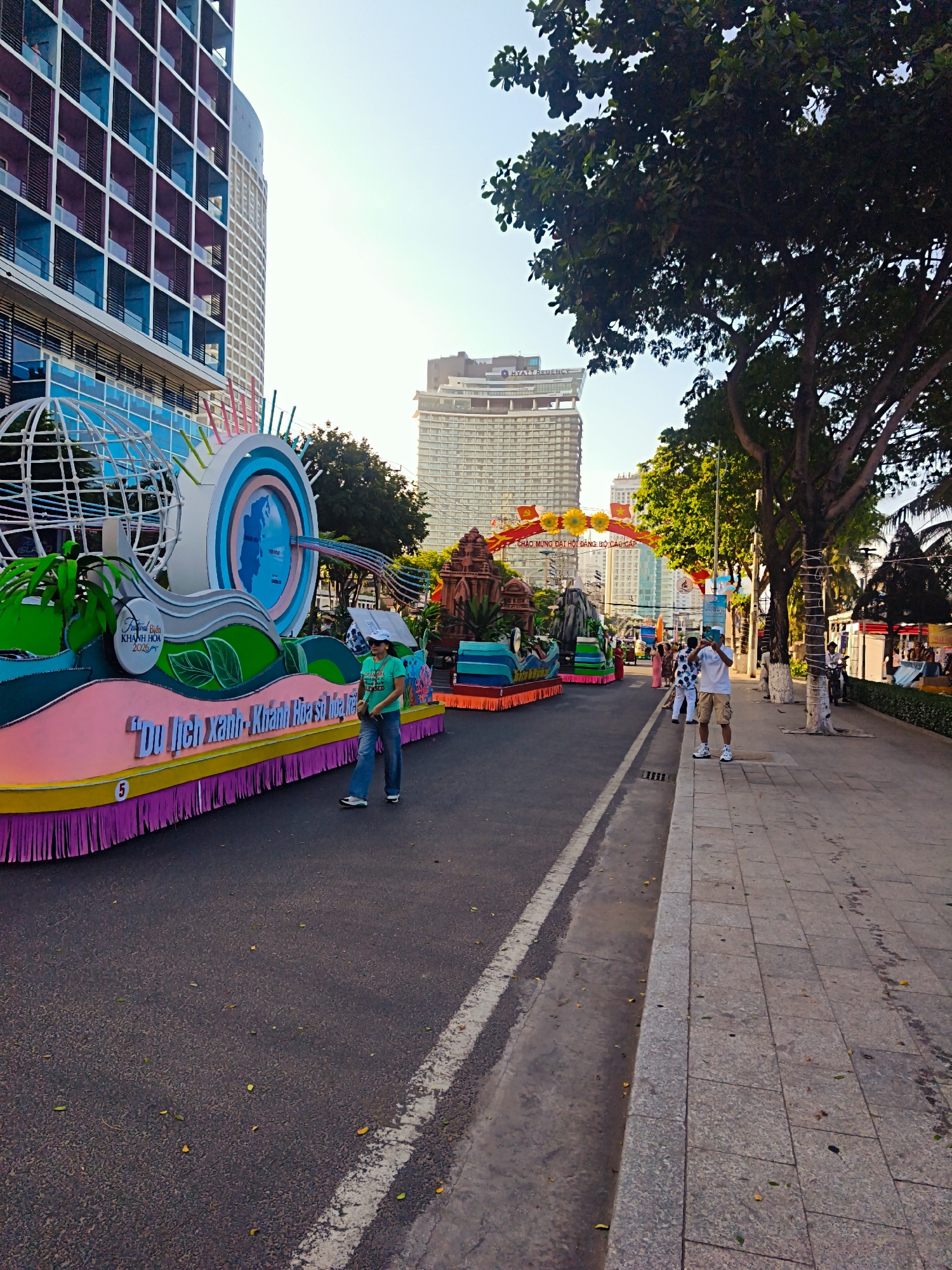The image captures a street scene in Nha Trang, Vietnam, likely during a festival or parade. In the foreground, a large, colorful float with a globe and a circular design is prominently displayed. The float is decorated with green foliage and text in Vietnamese. A woman in a green t-shirt and jeans is standing next to the float, looking towards the camera.
The street is paved with asphalt, and a white line marks the lane division. To the right of the street is a sidewalk made of stone tiles, with trees lining the edge. Several people are visible on the sidewalk, some standing and observing, while one man in a white shirt and khaki shorts is taking a picture with his phone.
In the background, modern buildings with distinctive architectural features rise against a clear sky. One building on the left is tall and features a facade with blue and purple grid patterns. Another building in the mid-ground is a large, lighter-colored structure with many windows. A festive archway with Vietnamese writing and yellow floral decorations spans the street in the background, suggesting a celebratory event. The overall atmosphere appears bright and sunny, with scattered leaves on the ground indicating an outdoor setting.