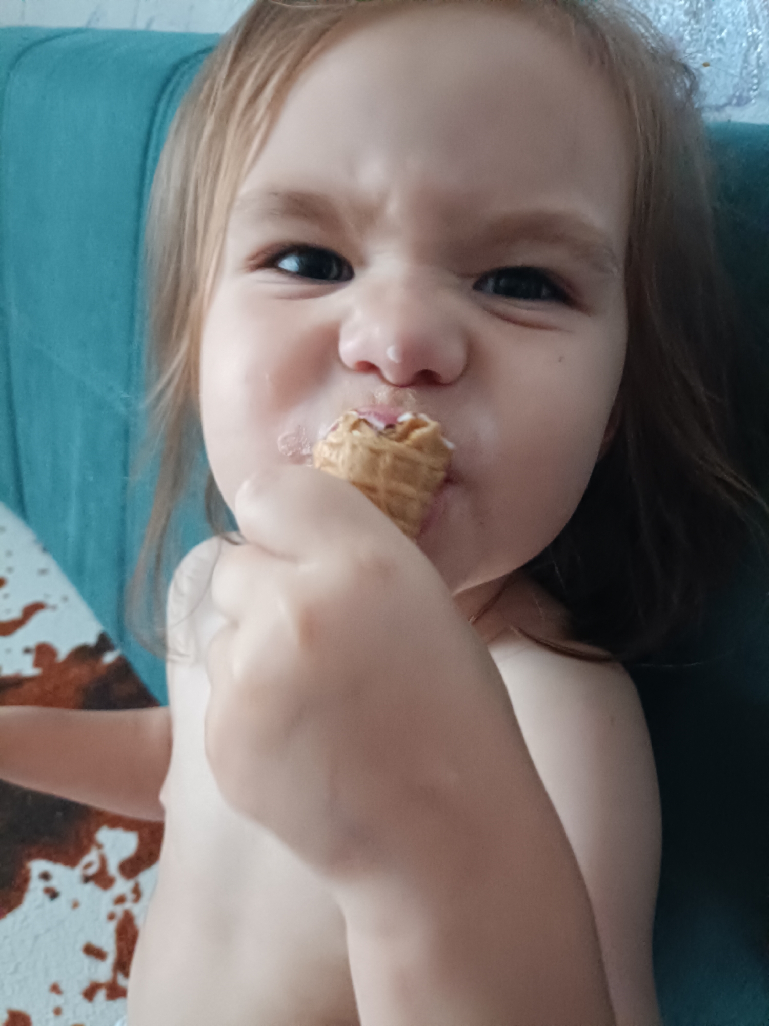 A close-up shot shows a young child, likely a toddler, with light brown hair and fair skin, enjoying an ice cream cone. The child's face is the primary focus, with eyes wide and a slight grimace as they eat, indicating intense concentration or perhaps a strong taste. The child is holding the ice cream cone in their right hand, bringing it to their mouth. Their torso is visible, and they appear to be topless. The background is softly blurred, with a hint of a teal-colored surface behind the child's head, possibly a chair or sofa. To the left, a patterned surface with brown and white splotches is visible, reminiscent of an animal hide print rug. The lighting suggests an indoor setting, possibly during the daytime, with natural light contributing to the illumination. The child's expression conveys a mix of focus and perhaps a touch of playful struggle with their treat. No text is visible in the image.