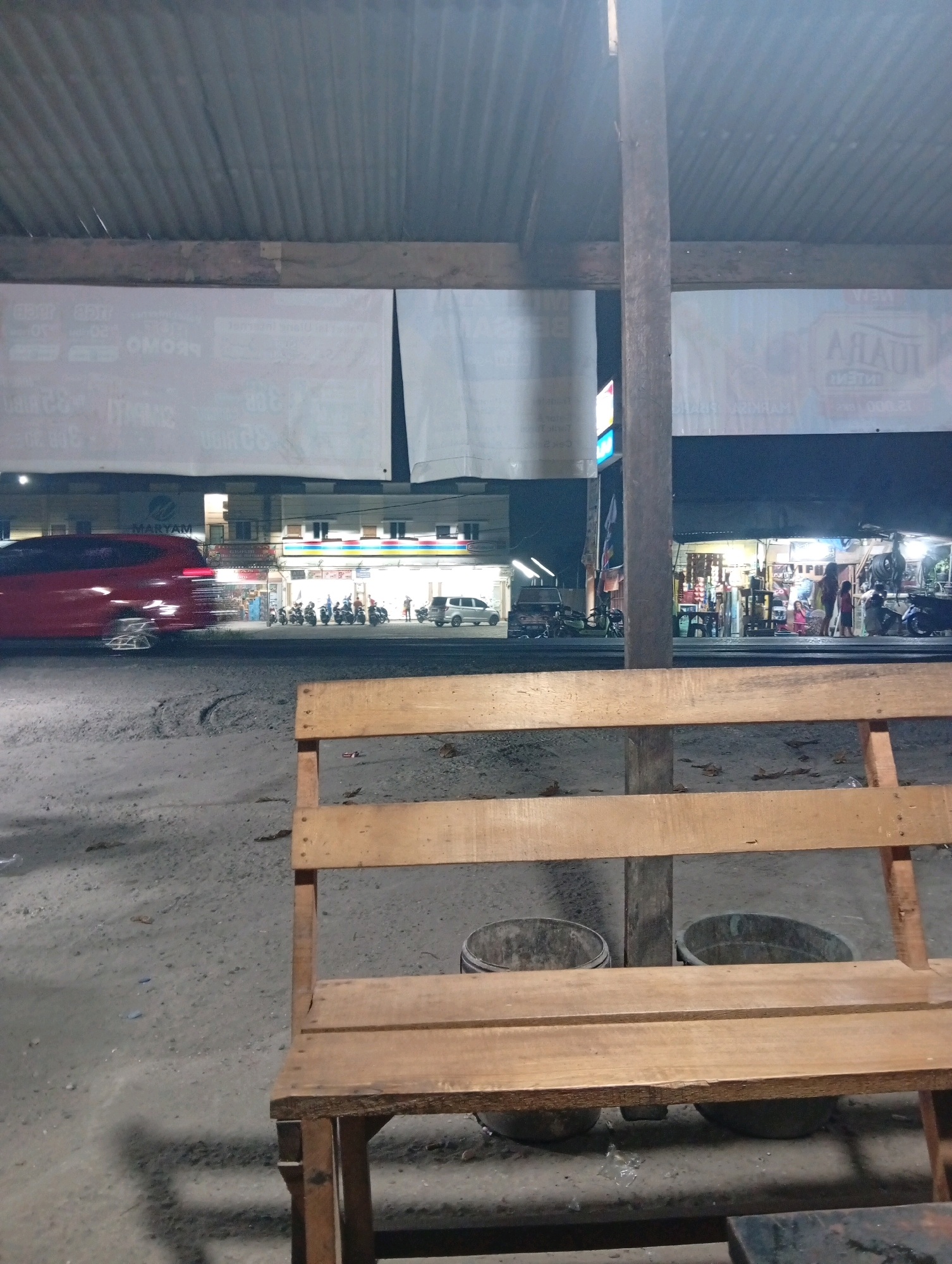 The image depicts an outdoor scene at night, likely a roadside area in Kubang Jaya, Indonesia. A wooden bench is in the foreground, facing away from the viewer. Behind the bench, the ground is a mixture of dirt and gravel. Two large, dark-colored buckets are visible on the ground behind the bench.
In the background, there are several buildings with illuminated storefronts. On the left side, a red car is moving, its motion blurred by the slow shutter speed. A row of parked motorcycles is visible in front of one of the buildings. People can be seen standing or walking in front of the shops. The overall impression is that of a busy commercial street at night. The sky is dark, and the lighting comes primarily from the artificial lights of the shops and streetlights. A corrugated metal roof is supported by wooden beams and a central wooden pillar. Large white banners are hung across the top of the structures.