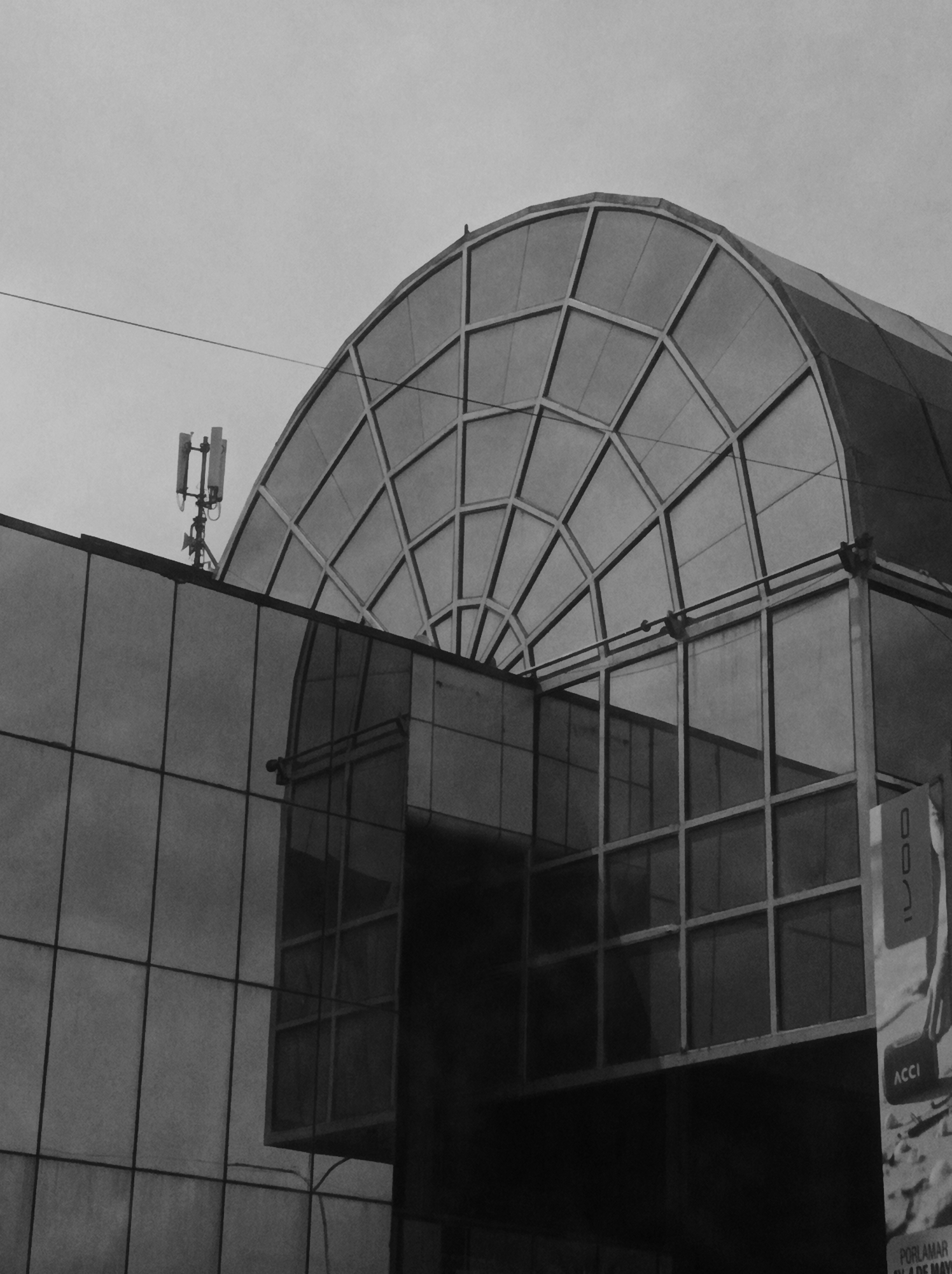This black and white photograph depicts a modern building characterized by its prominent arched glass and metal roof structure. The arch, composed of numerous rectangular and trapezoidal glass panes framed by a grid, curves gracefully across the upper part of the image. Below this, the building's facade is dominated by large, rectangular glass panels, reflecting parts of the structure itself and the overcast sky.
To the left, a telecommunications antenna, featuring multiple vertical components, stands atop a flat section of the building. A thin, horizontal wire or cable traverses the upper left portion of the frame. On the far right, a vertical poster or banner is partially visible. Clearly discernible text on the poster includes "ACCI" and, further down, "PORLAMAR" and "EL 4 DE MAYO". Above "ACCI", the characters "00 / I" (or "00 / 1") are also present.
The scene suggests an urban or commercial setting, showcasing modern architecture. The uniform grey of the sky indicates an overcast day, contributing to the photograph's subdued, monochromatic atmosphere. No specific activity or individuals are depicted, focusing instead on the geometric forms and reflective surfaces of the architectural elements. The visible text confirms the location as Porlamar, Venezuela.