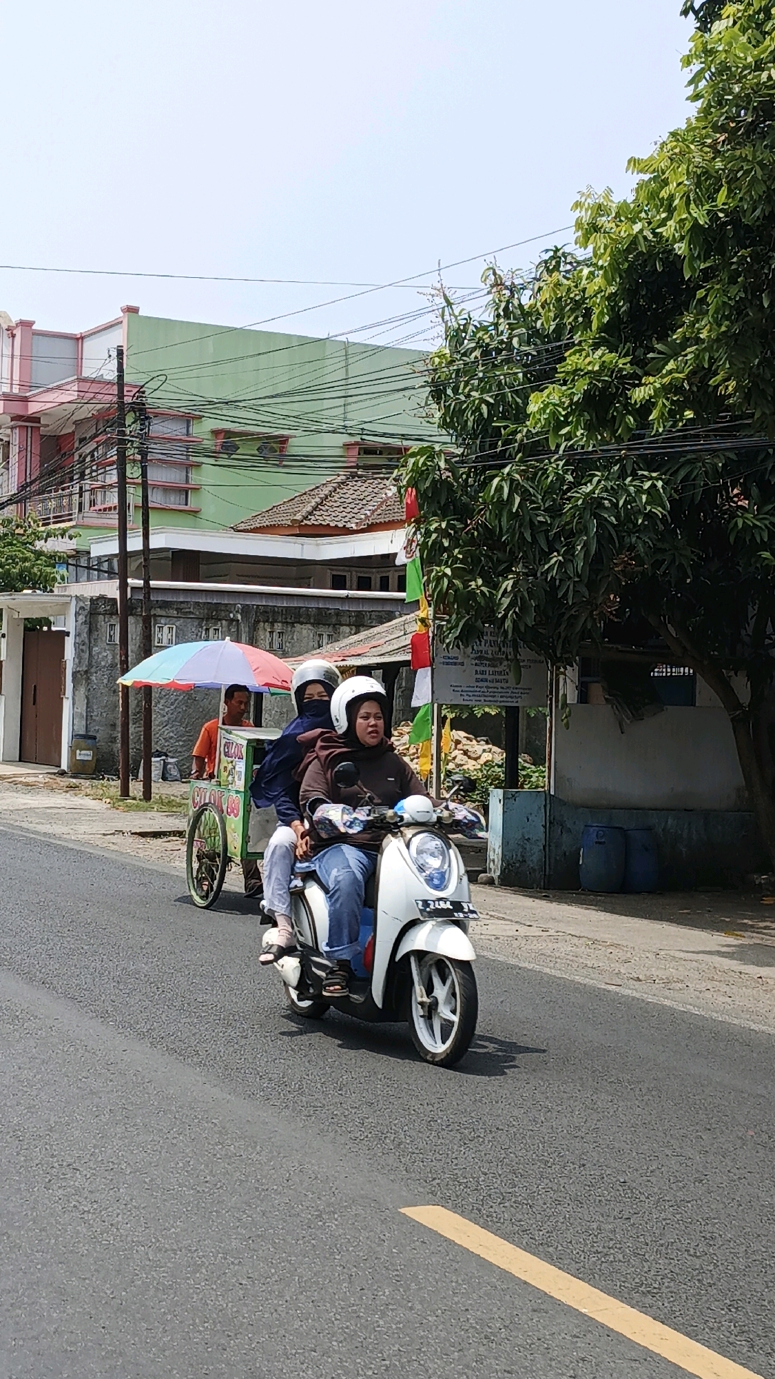 A three-wheeled motorcycle with a canopy is driving on a paved road in Cikembulan, Indonesia. The motorcycle is white and has three wheels, with a passenger sitting behind the driver. A child is sitting in the sidecar of the motorcycle, and a colorful umbrella is covering the child. The motorcycle is decorated with flags and banners. The driver and passenger are wearing helmets. The background of the image shows a building with a green roof and a pink facade, and a large tree with green leaves. The road has a yellow line painted in the center.
