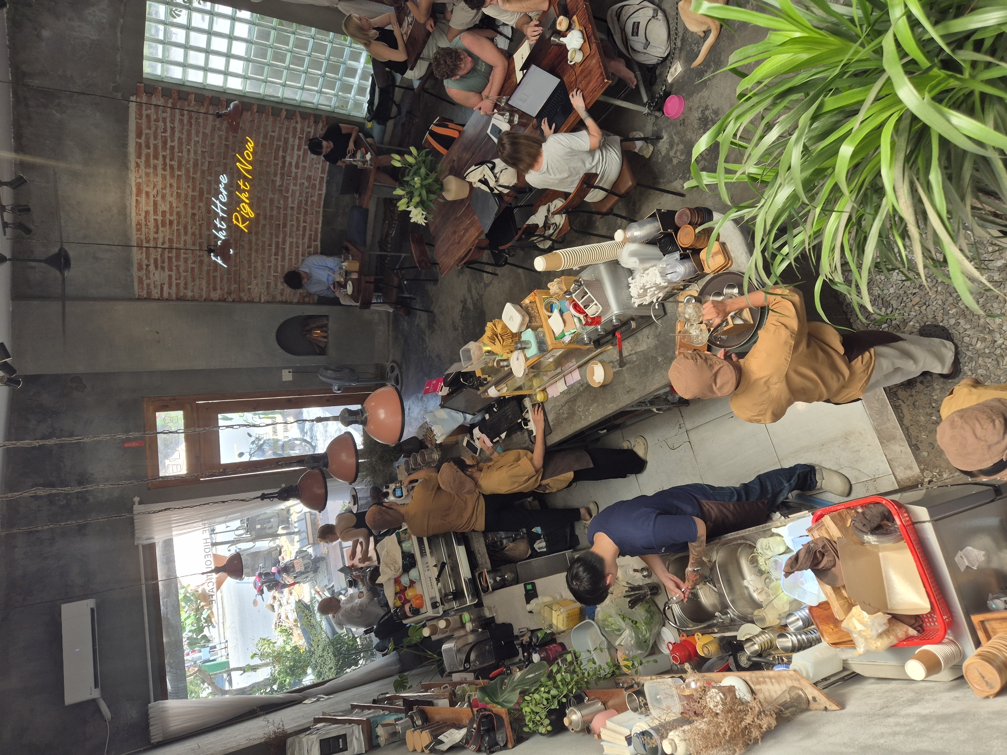 This is an interior, high-angle shot of a busy coffee shop in Da Nang, Vietnam. The space has a raw, industrial aesthetic with concrete walls and ceiling, exposed brick on one wall, and a large glass block window. Several people are present, including baristas working behind a counter and customers seated at tables.
In the foreground, to the left, a male barista in a blue shirt and brown apron is washing dishes in a double sink. To his right, a female barista in a tan uniform and apron is operating a coffee machine and interacting with a customer. Further back, another barista in a tan uniform is also behind the counter.
The cafe is filled with wooden tables and chairs, some occupied by customers working on laptops or talking. A neon sign on the brick wall reads "F*ck Here Right Now". Plants are interspersed throughout the space, adding a touch of greenery. Through a large window on the left, a street scene with parked motorcycles and greenery is visible, suggesting the cafe is located on a street.
The overall impression is a vibrant, modern cafe in Vietnam, bustling with activity.