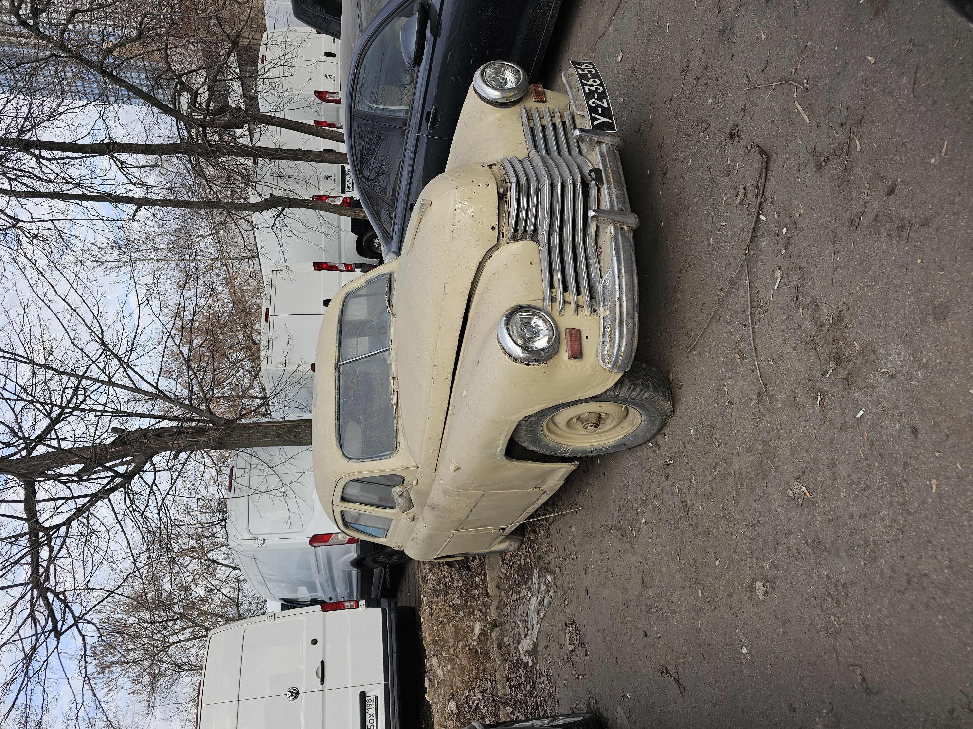 The image depicts a weathered, vintage car parked in an outdoor location, likely in the city of Khimki, Russia, as indicated by the license plates seen on some vehicles in the background. The old car has a beige exterior and shows signs of disrepair. The car is situated next to a black modern car, and a row of white vans. Bare trees are visible in the background, suggesting the photo was taken in a temperate climate during a colder season. The ground is pavement. There are no people visible.