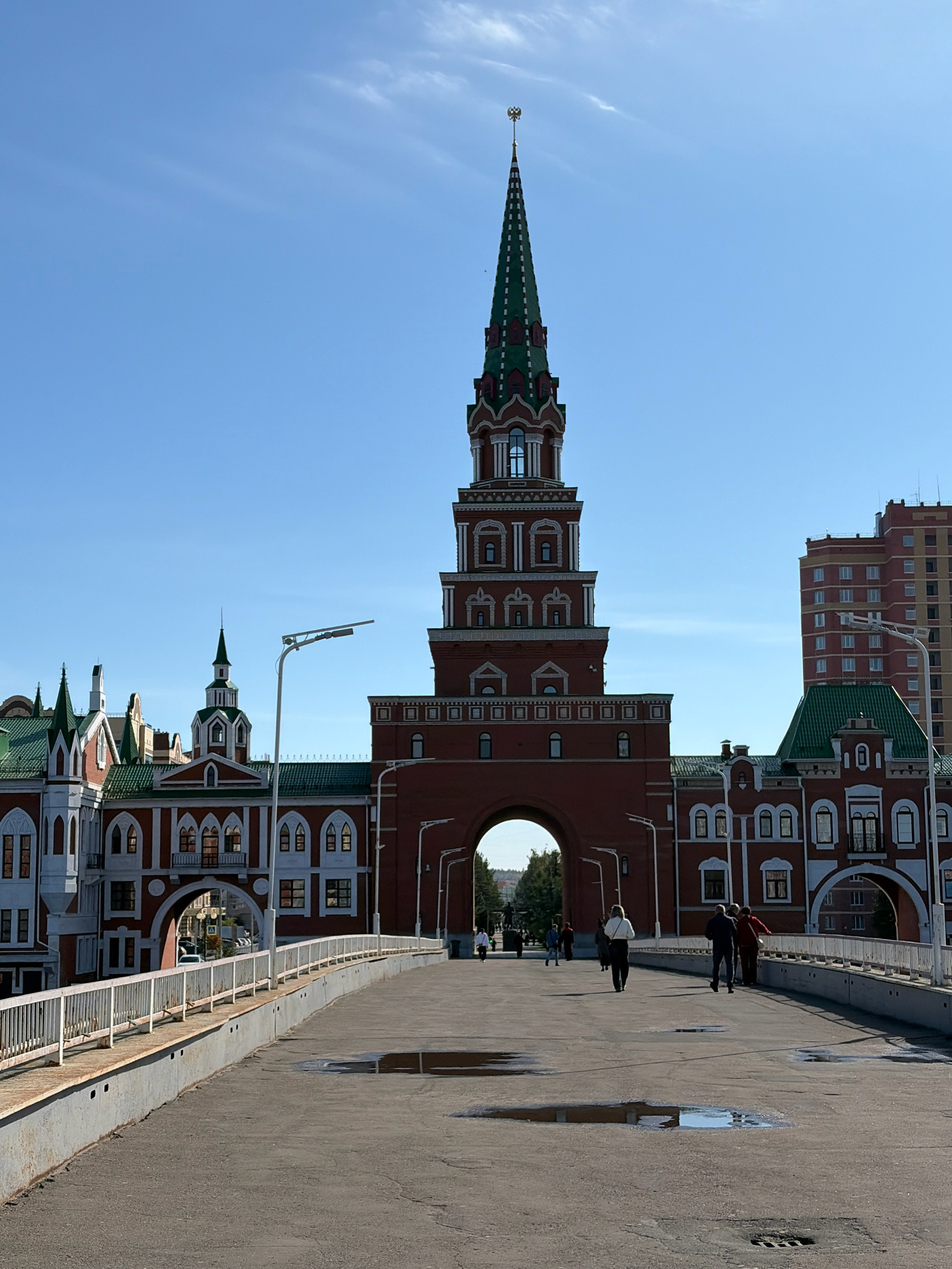 The image captures a view of the National Art Gallery building in Yoshkar-Ola, Russia. The foreground features a paved pedestrian bridge with a white metal railing on the left side. Puddles of water are visible on the bridge's surface. Several individuals are walking on the bridge.
The central focus of the image is the distinctive architecture of the National Art Gallery, which resembles a red brick Kremlin-style tower with a tall green spire topped with a golden emblem. The building exhibits ornate architectural details, including arched entrances and windows, and is flanked by other structures with similar stylistic elements, characterized by red brick facades and green roofs.
In the background, a modern, multi-story residential building with red and yellow brickwork stands to the right of the National Art Gallery. The sky is clear and blue with a few wispy clouds. The overall scene suggests a sunny day in an urban environment, highlighting a prominent landmark in the city.