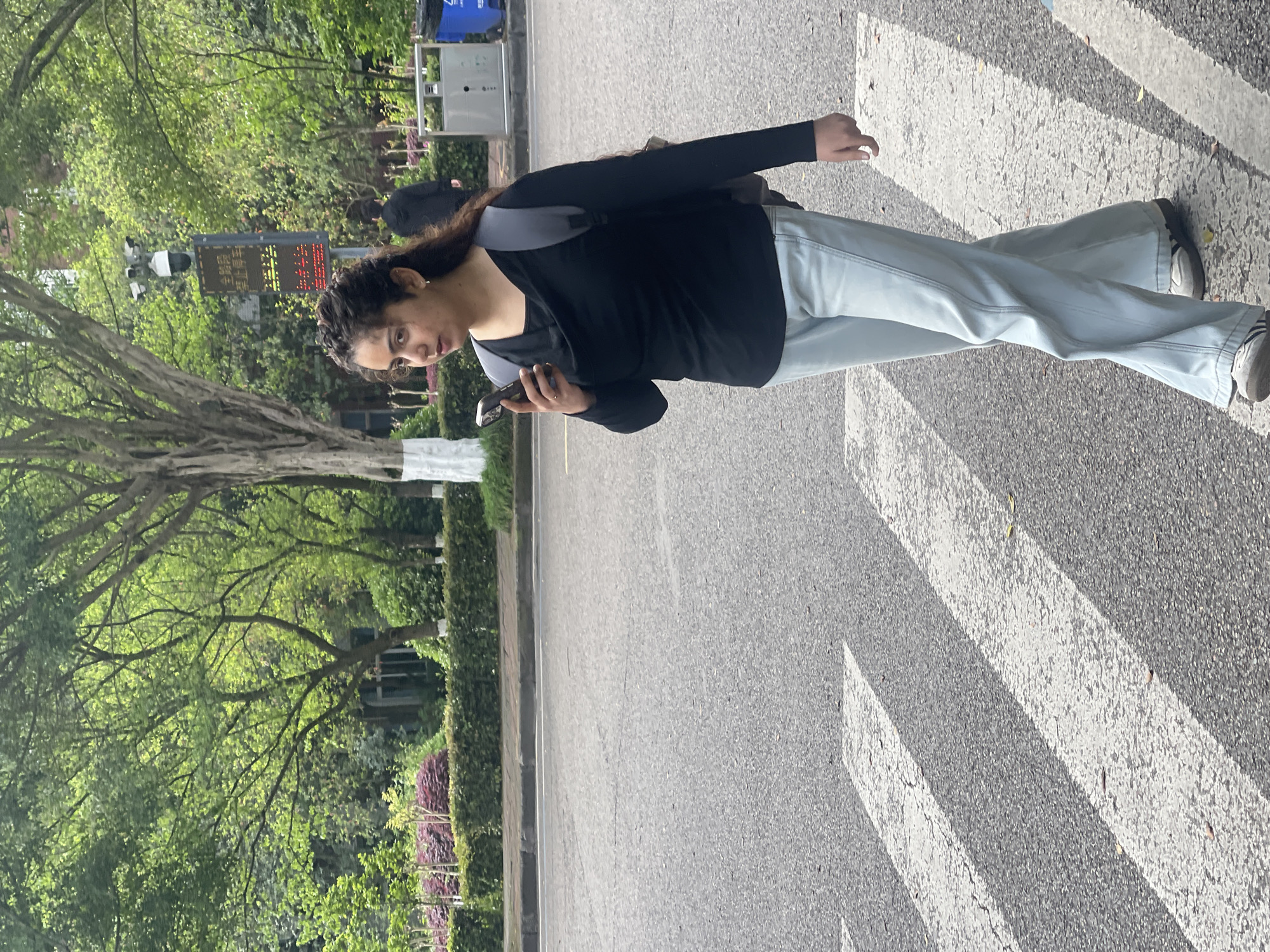 A woman stands on a crosswalk in what appears to be a streetscape in Chongqing High-tech Zone, China. She has dark hair and wears a black top, light-colored pants, and a backpack with gray straps. She is holding a smartphone. The crosswalk is white against the gray asphalt. To her left, there is a green area with trees and shrubs. A street sign is visible in the background.