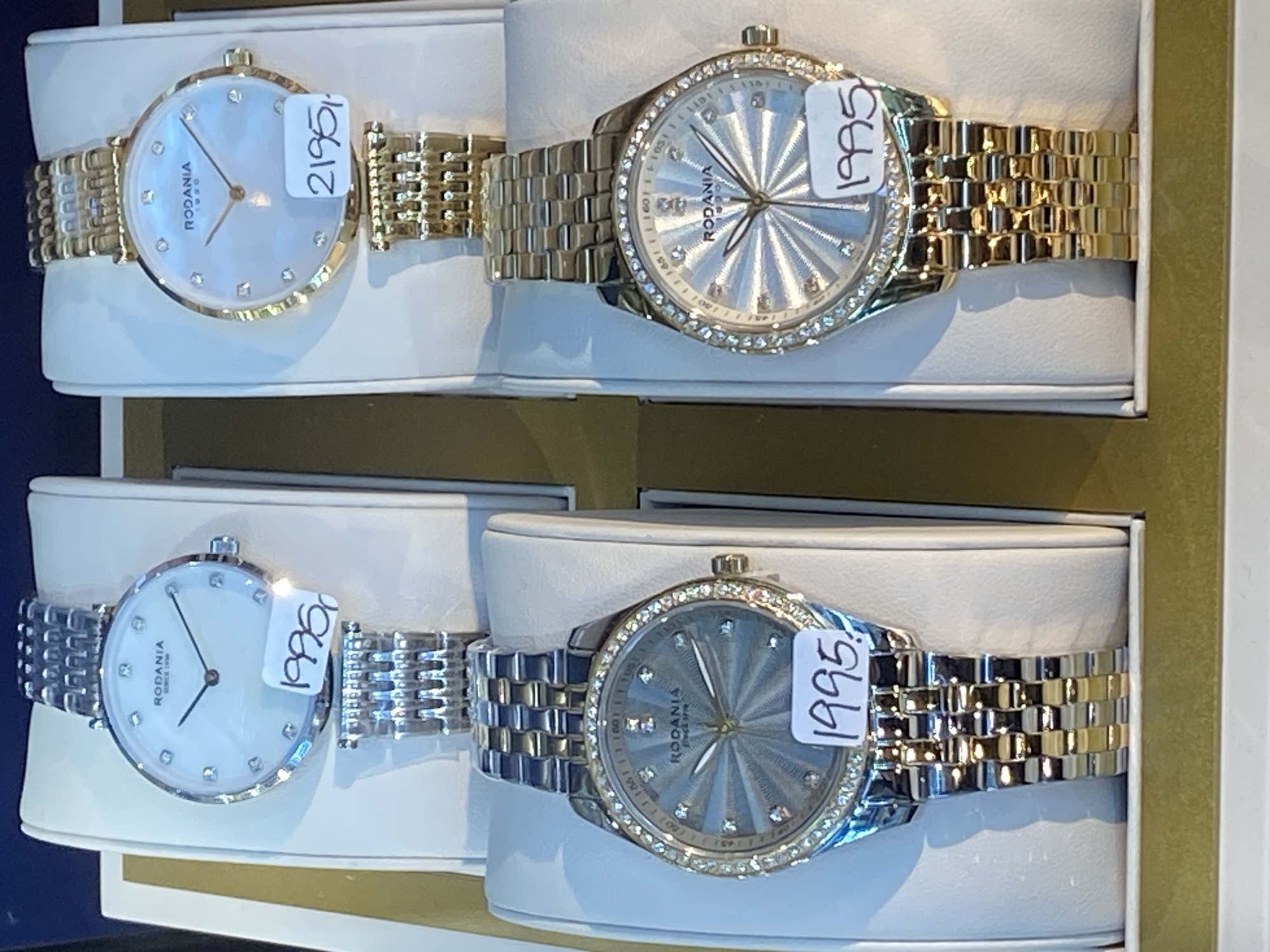 The image shows a display of four watches in a retail setting, possibly a jewelry store. The watches are arranged in pairs, with two watches on the top row and two on the bottom row. All watches appear to be of a similar style, with round faces and metal bands. The top row features two watches with silver-toned bands and white faces, one with a price tag of "1995" and the other with a price tag of "2195". The bottom row displays two watches, both with diamond-encrusted bezels. The watch on the left has a silver and gold-toned band and a blue-grey face with a price tag of "1995". The watch on the right has a gold-toned band and a white face, with a price tag of "1995". The watches are presented on white display stands within a box-like structure. The overall setting suggests a commercial environment. The location of Herning, Denmark is not discernible from the image.