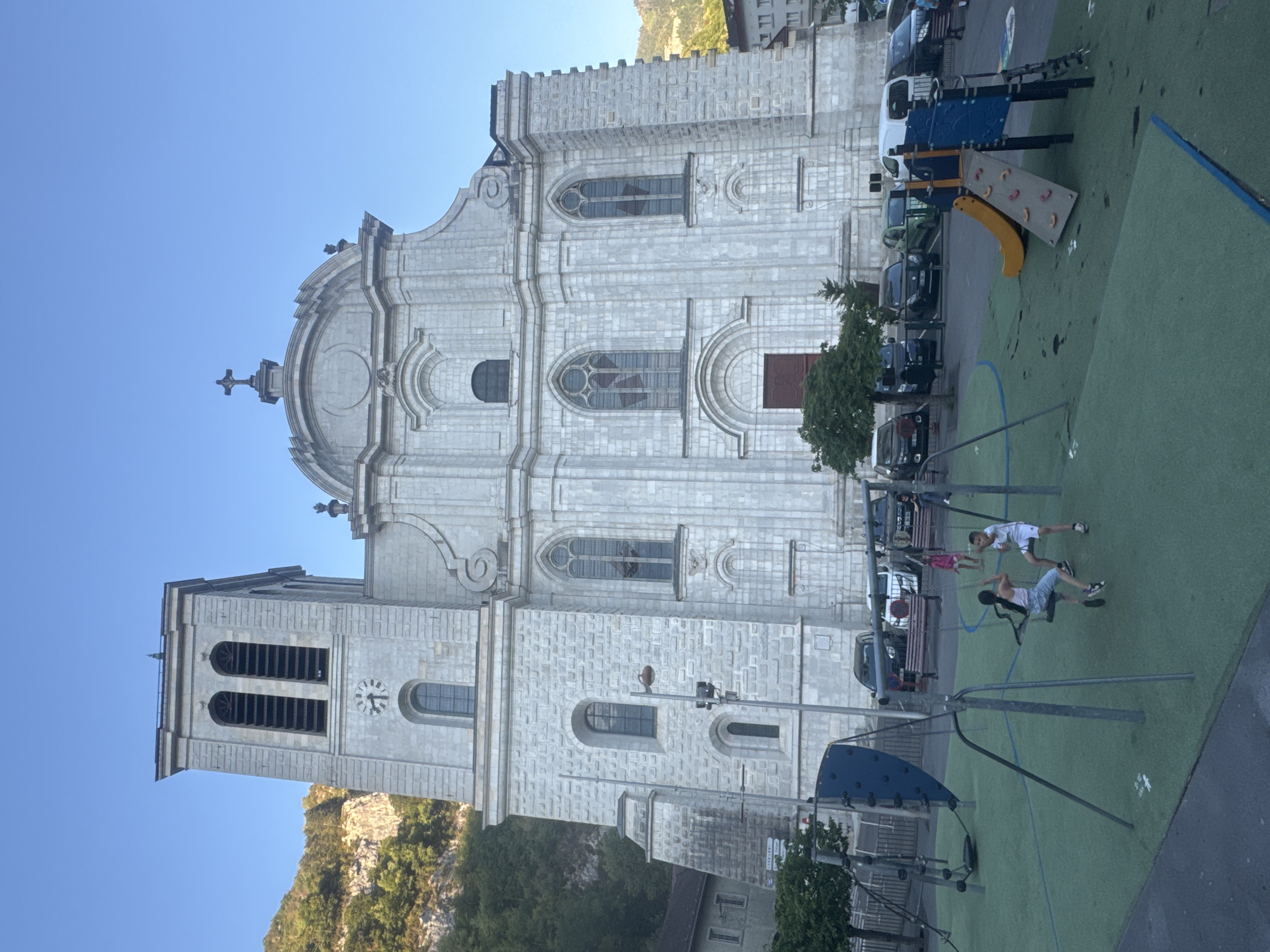 The image shows a large, white stone church with a tall bell tower on the left. The church has a classic facade with arched windows and decorative elements. In front of the church is a playground with swings and a slide. Several cars are parked in the background. Two children are on swings in the foreground, and other people are visible in the background near the cars and on the playground. The scene appears to be in a town or city, possibly Saint-Claude, France, given the architectural style and the presence of a church with a bell tower, common in European towns. The sky is clear and blue, indicating it is daytime.