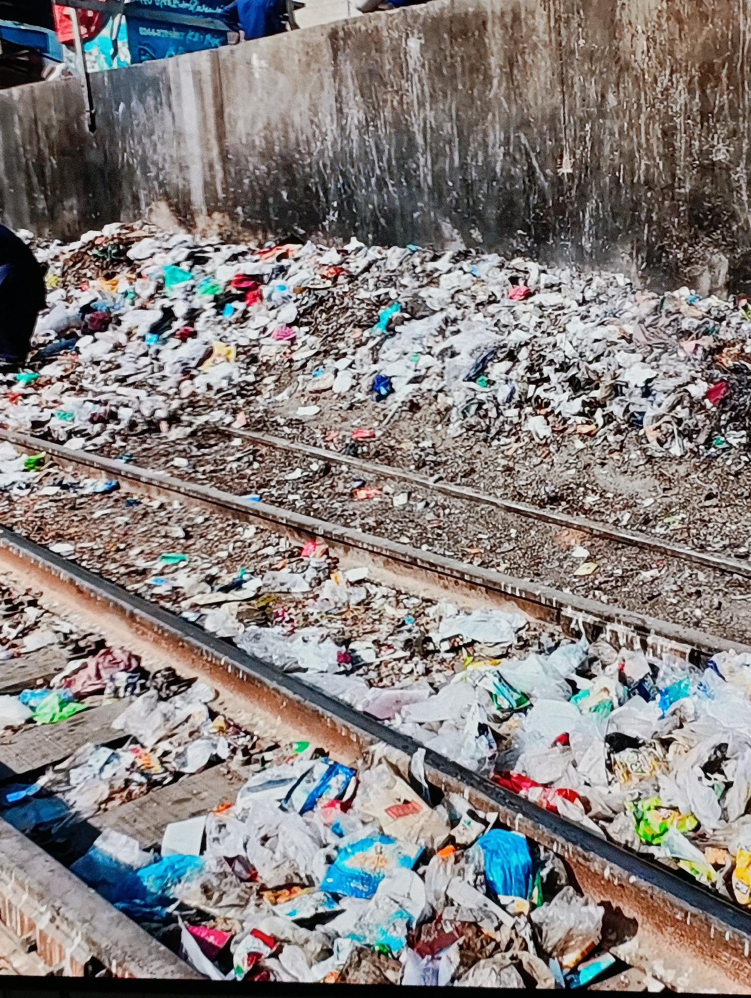 The image shows a large pile of garbage accumulated on and around a set of railway tracks. The garbage appears to consist primarily of plastic bags and wrappers, with a mix of colors visible. A concrete wall serves as a backdrop to the garbage pile. The location appears to be in an urban area, but the specific city and country are not identifiable from the image.