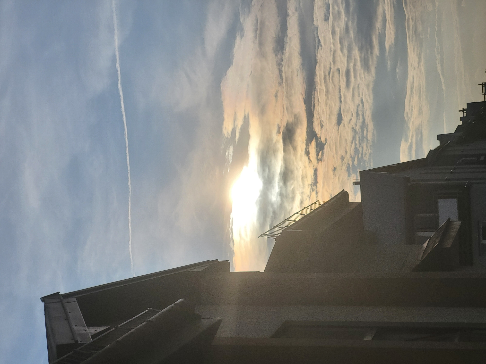 A photograph taken from a low angle shows the edge of a building silhouetted against a sky with scattered clouds. The sun is visible, partially obscured by clouds, creating a bright, luminous effect with a rainbow halo. A thin contrail from an aircraft stretches horizontally across the sky. The building appears to be residential, with multiple stories and windows visible. Another building, with a pitched roof, is partially visible in the foreground. The sky is a mix of light blue and white, with the sun casting a warm glow on the clouds.