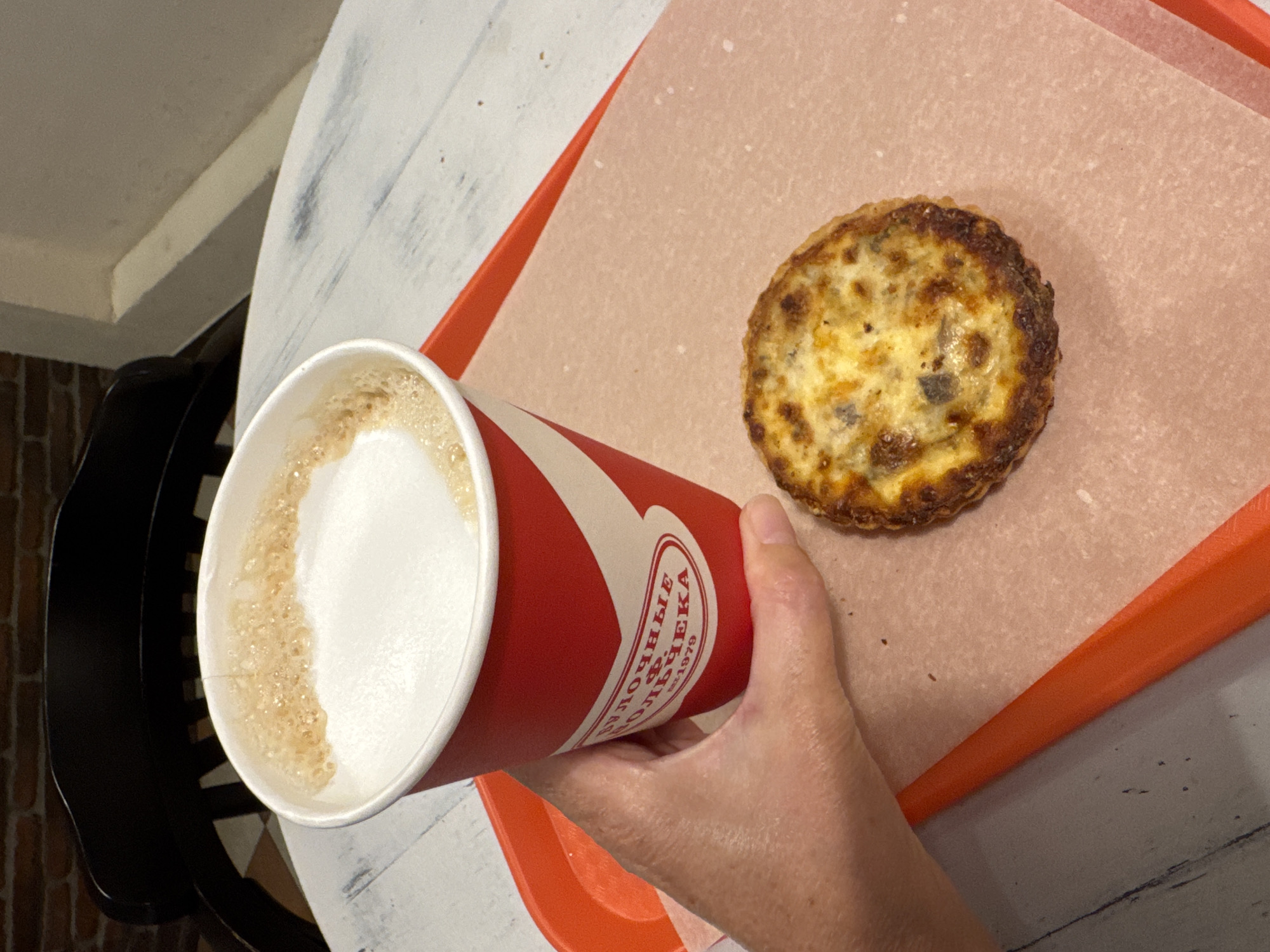 A hand holds a red and white paper cup filled with a frothy beverage, likely coffee or latte. The cup has text printed on it, partially visible, including "ВОЛШЕБНЫЕ" and "БУЛОЧЕК", suggesting a bakery or pastry shop. Next to the cup, on an orange tray lined with parchment paper, sits a small, round quiche with a golden-brown crust and visible filling. The background shows a white wooden table with some scuff marks and a hint of a dark chair and brick wall, indicating an indoor setting. The lighting appears to be natural, possibly daylight, suggesting it's daytime. The scene evokes a casual breakfast or snack moment, perhaps at a cafe in Vsevolozhsk, Russia, given the location context and the Cyrillic text on the cup. There are no strong emotional cues, but the overall impression is one of a simple, everyday enjoyment.