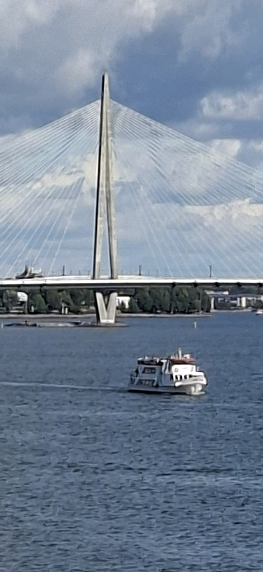 A cable-stayed bridge spans a body of water under a partly cloudy sky. A white and red boat is moving on the water in the foreground. The bridge has a tall, triangular pylon with many cables extending from it to the bridge deck. Trees and buildings are visible on the far bank. The scene is consistent with the Baana bridge in Helsinki, Finland.