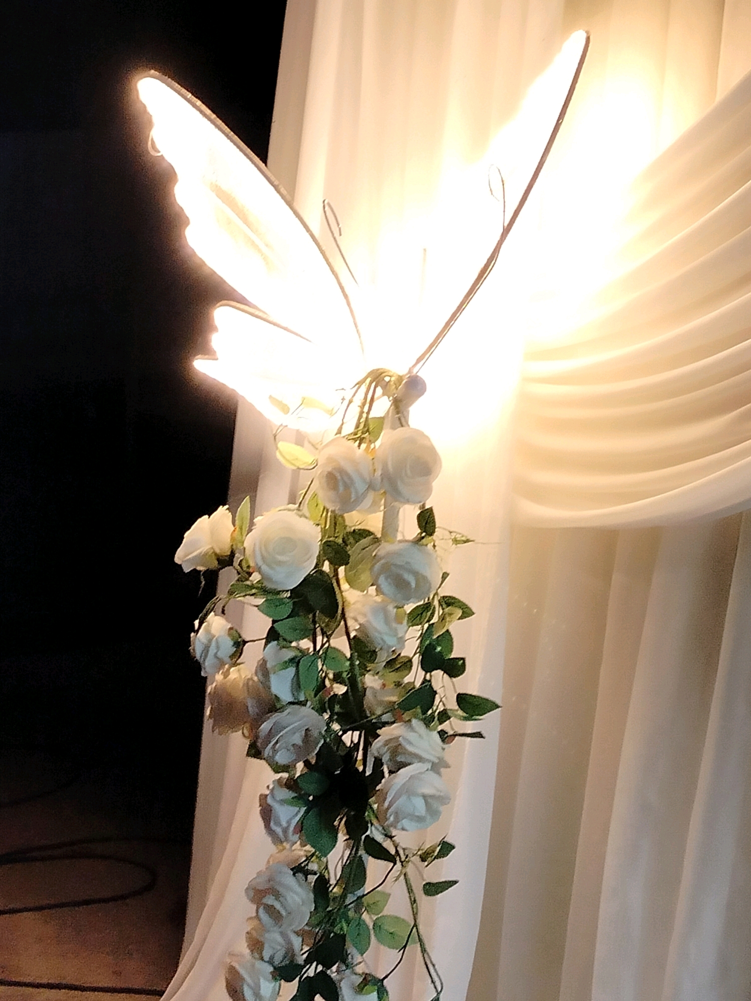 A backlit butterfly-shaped decorative object is the central focus, with its wings glowing brightly against a dark background on the left and draped, cream-colored fabric on the right. A cascade of artificial white roses with green leaves hangs from the bottom of the butterfly. The scene appears to be an indoor setting, possibly a venue or event space. There are no people present in the image. The location of Көлкент, Kazakhstan, is not explicitly visible or suggested by any elements within the image itself.