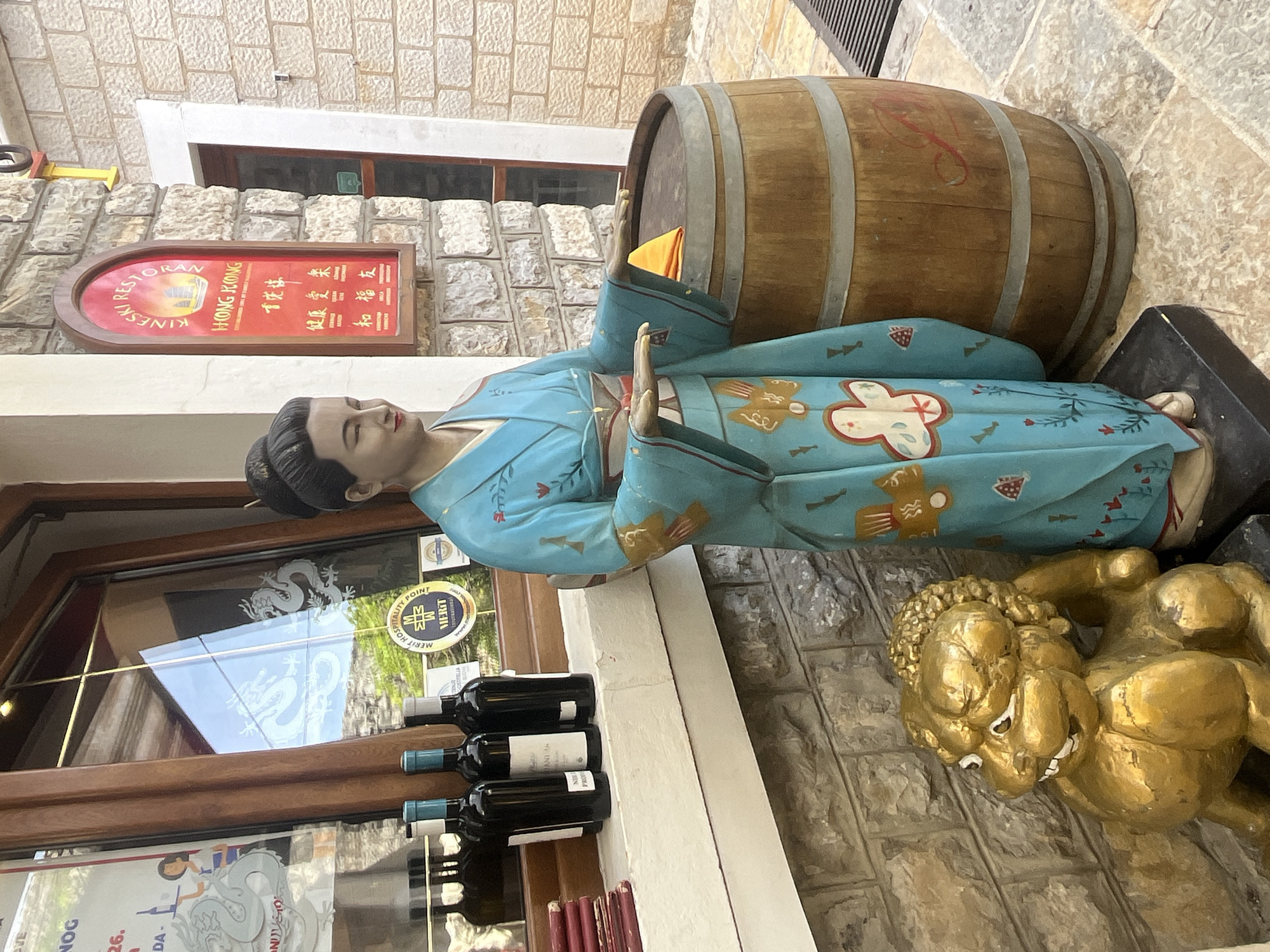 The image shows a storefront in what appears to be a European-style city. The statue of a Japanese geisha is in the foreground. Next to the geisha is a wooden barrel. Next to the geisha is a golden lion statue. Behind the geisha are wine bottles on a white shelf. The glass door behind the wine bottles has a white dragon decal. Next to the geisha is a sign in red and black for a restaurant called "Hong Kong." The storefront is located on a street paved with stone.