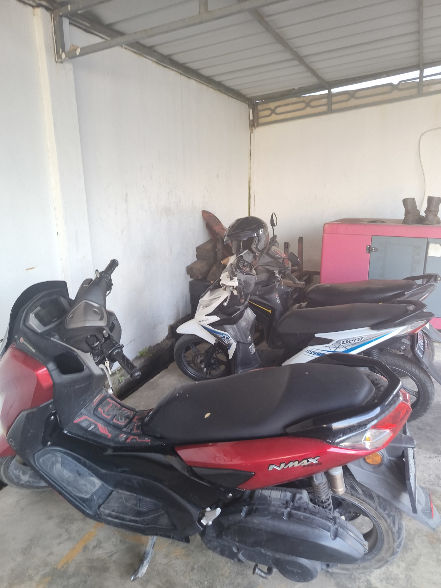 A red and black scooter with a "NMAX" logo is parked in front of other motorcycles and a pink generator. The scene appears to be an outdoor covered area with a corrugated metal roof supported by metal beams. The walls are white and the ground is concrete with yellow markings. Two pairs of dark boots are placed on top of the pink generator.