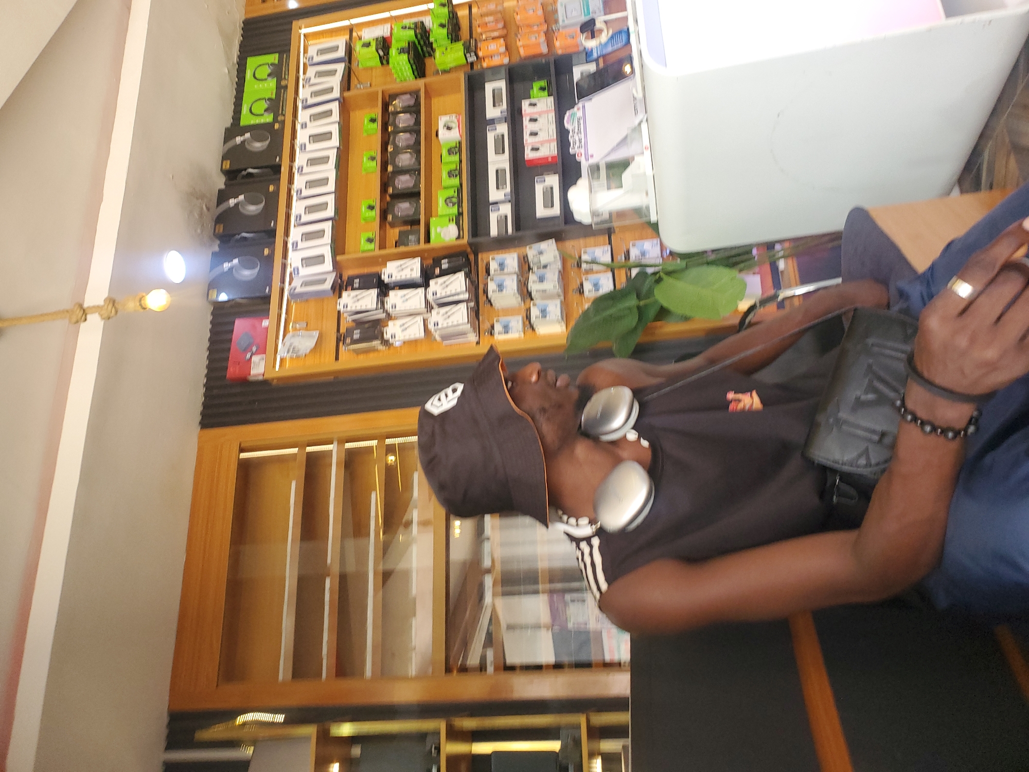 A Black male wearing a black bucket hat and a black tank top with white stripes on the sleeves is sitting on a black bench. He has silver headphones around his neck. He is also wearing a black wristband and a gold ring on his right hand. A black fanny pack is slung across his chest. The man is in a retail store with shelves of electronic products behind him. The shelves are stocked with various items including boxed headphones, phone cases, and charging cables. The store appears to be well-lit, with a bright light fixture visible at the top of the image. The overall scene suggests a commercial environment, possibly a mobile phone or electronics store in Jos, Nigeria, given the context.