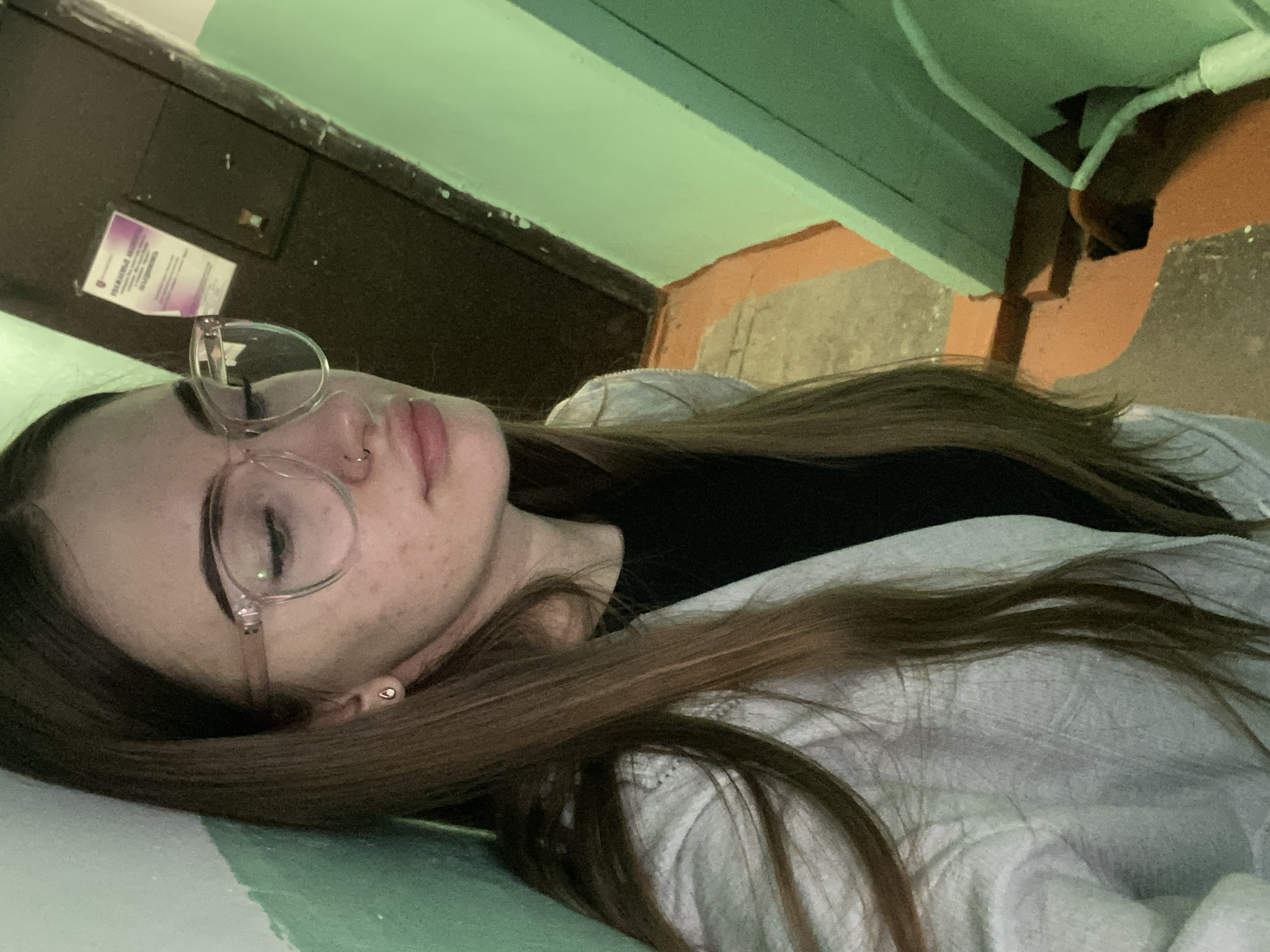 A young female with long, straight, brown hair, wearing clear-framed glasses, a small hoop nose ring in her left nostril, and a stud earring in her left ear, is depicted in an indoor setting. Her eyes are directed downwards, and her lips are slightly parted. She is dressed in a black top layered under a light-colored, possibly white or light grey, long-sleeved shirt or jacket. The subject is positioned against a bright green painted wall. The background shows a continuation of the green wall, a dark brown/black door or panel with a light purple paper notice containing non-English text affixed to it, and a section of concrete or similarly textured floor with patches of orange-brown coloration. A white cable or pipe is visible along the lower right wall-to-floor junction. The location appears to be an interior hallway or common area of an older building.