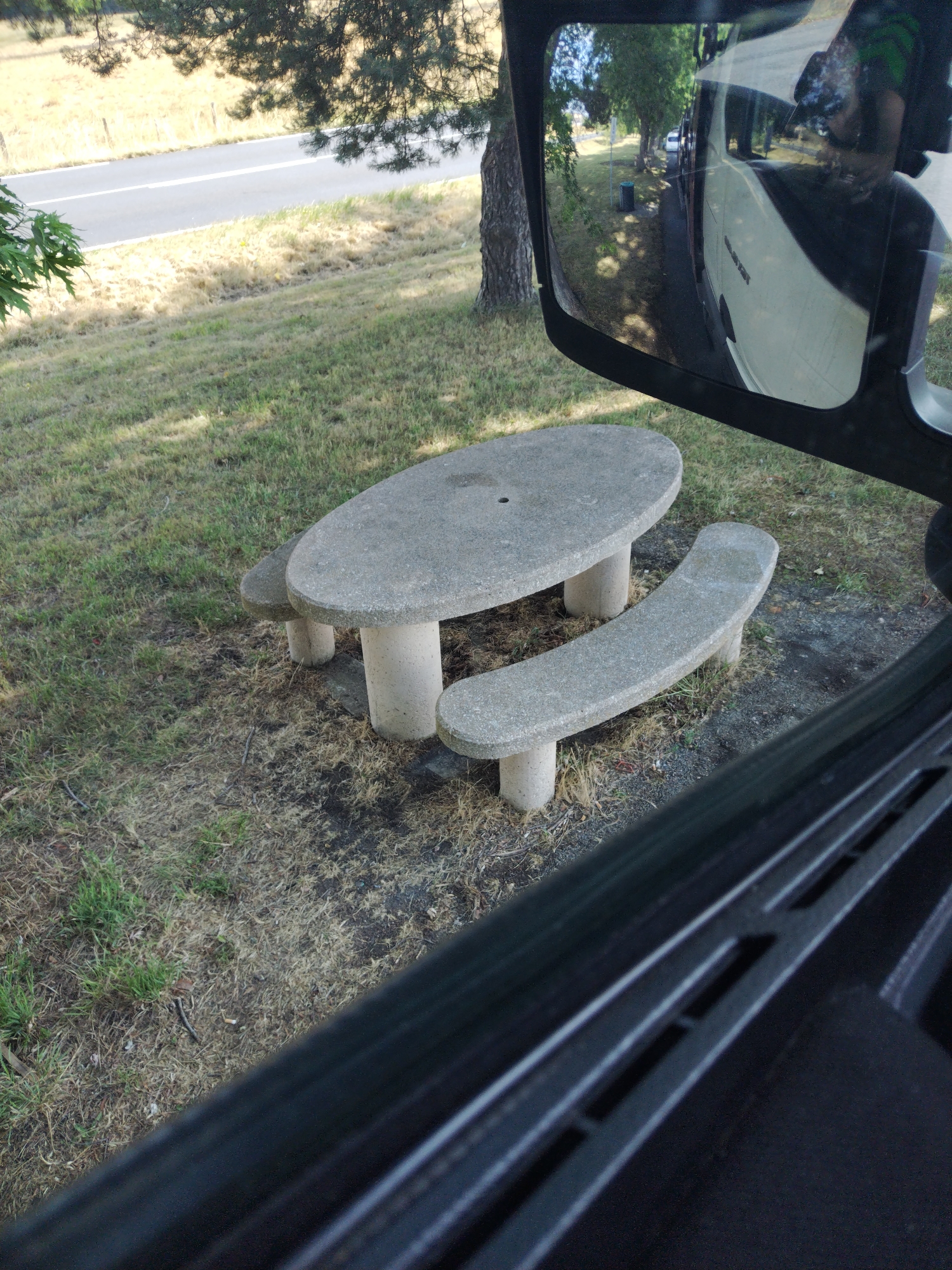 This media file shows a stone picnic table with attached benches in an outdoor setting. The table is oval-shaped and supported by cylindrical legs. Two benches, also made of stone, are attached to the table, one curved and the other more straight. The picnic table is situated on a grassy area with dry patches and scattered twigs. In the background, a paved road is visible, bordered by dry, yellowed grass. Trees are present, casting shadows on the ground. A reflection in what appears to be a mirror or window shows a white vehicle, possibly a truck, and trees lining a road. The lighting suggests it is daytime, likely with bright sunlight given the shadows. The overall scene appears to be a rest stop or picnic area along a road in Magnet, France. There are no people visible in the scene.