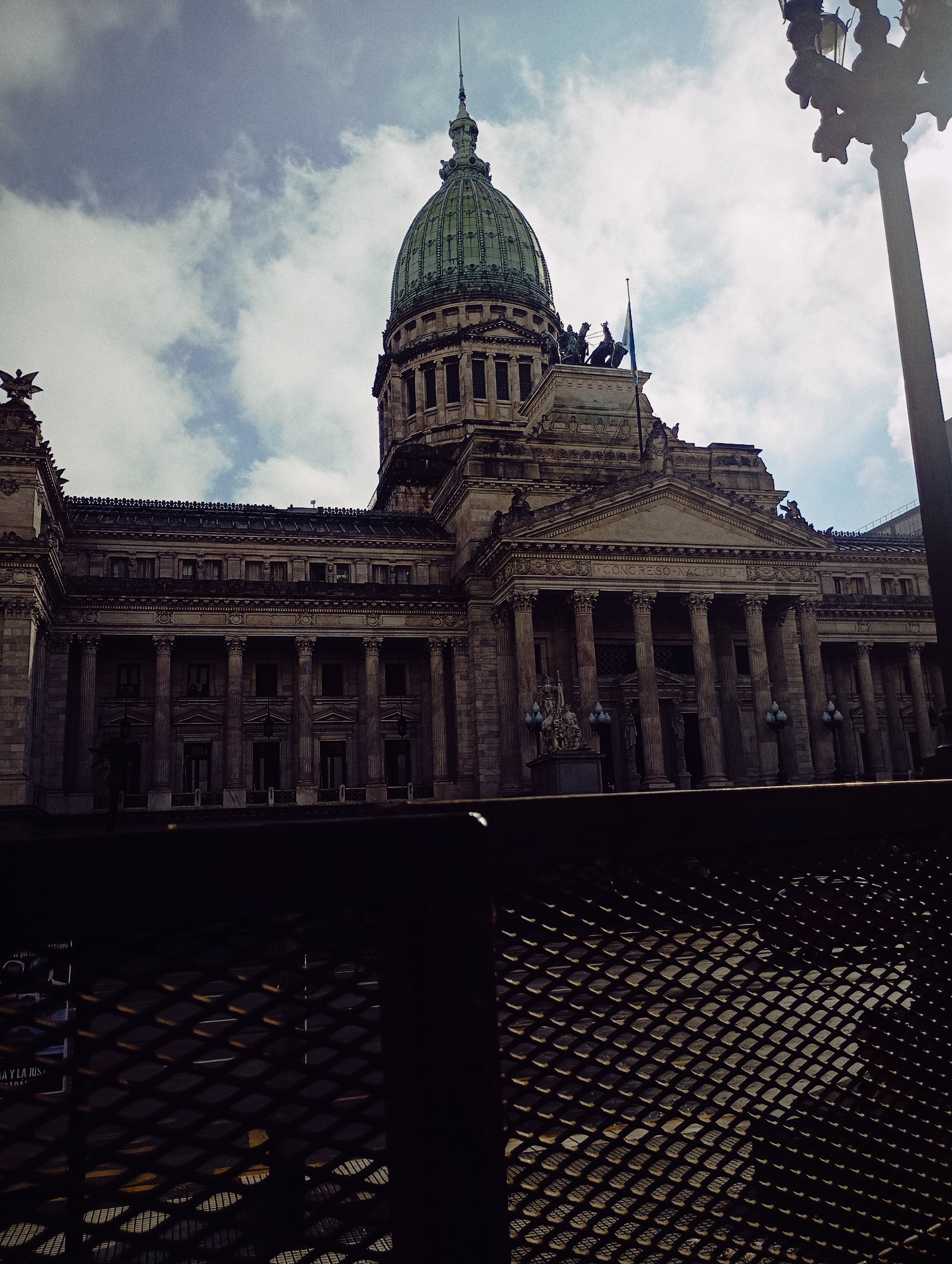 The image displays a grand, classical-style building, which is the National Congress (Congreso Nacional) of Argentina, located in Buenos Aires. The building is captured from a slightly low angle, partially obscured by a dark, diamond-patterned metal grate in the foreground.
The most prominent architectural feature is the large, green-patinated dome topped with a spired finial, rising majestically against an overcast sky. The main facade of the building features a monumental portico with numerous tall columns supporting a triangular pediment. Within the pediment, sculpted figures are visible, along with the text "CONGRESO NACIONAL." Above the pediment, on the roofline, there are additional sculptures and a flagpole flying a flag. To the far left, an ornate eagle-like sculpture adorns a corner of the building's roof.
The scene is set outdoors during the daytime, under a sky with scattered grey and white clouds. To the far right, a tall, multi-armed lamppost or street light fixture is partially visible, silhouetted against the sky. The overall lighting is somewhat subdued due to the cloudy conditions. No people are clearly visible, and no specific activity is taking place within the frame.