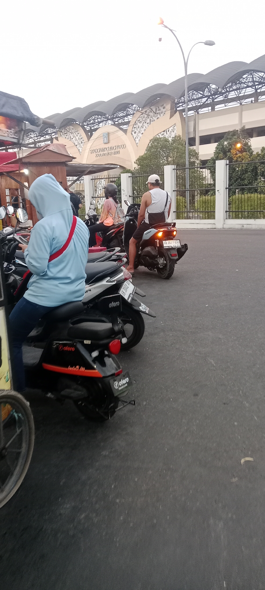 The image shows a street scene in Ngemplak, Indonesia, with a prominent stadium in the background. Several motorcycles are parked or stopped, and people are interacting with them.
In the foreground, on the left, a person wearing a light blue hooded sweatshirt and dark pants is seated on a motorcycle. They are looking down at their phone. A red strap is visible across their back. The motorcycle has a yellow and black color scheme.
To the right of this person, another motorcycle is parked, with a "o'fero" logo visible on its rear.
Further back, a motorcycle is occupied by two individuals. The person in the back is wearing a light orange and pink gradient shirt and a black helmet. They have a backpack with a pink strap. The person in the front is wearing a white tank top and a white cap, and is looking forward. This motorcycle is a dark color and has a license plate that reads "AB 3577 EAG". The motorcycle's brake lights are illuminated.
Behind the motorcycles, a fence with white posts and black railing runs across the scene. Beyond the fence is a large, modern stadium structure with a distinctive arched roof and decorative facade. Text on the stadium reads "STADION MAGUCHARLO KABUPATEN SLEMAN". Trees are visible near the stadium. A street light with a yellow light is also visible on the right side of the image. The ground is paved asphalt. The sky is overcast.