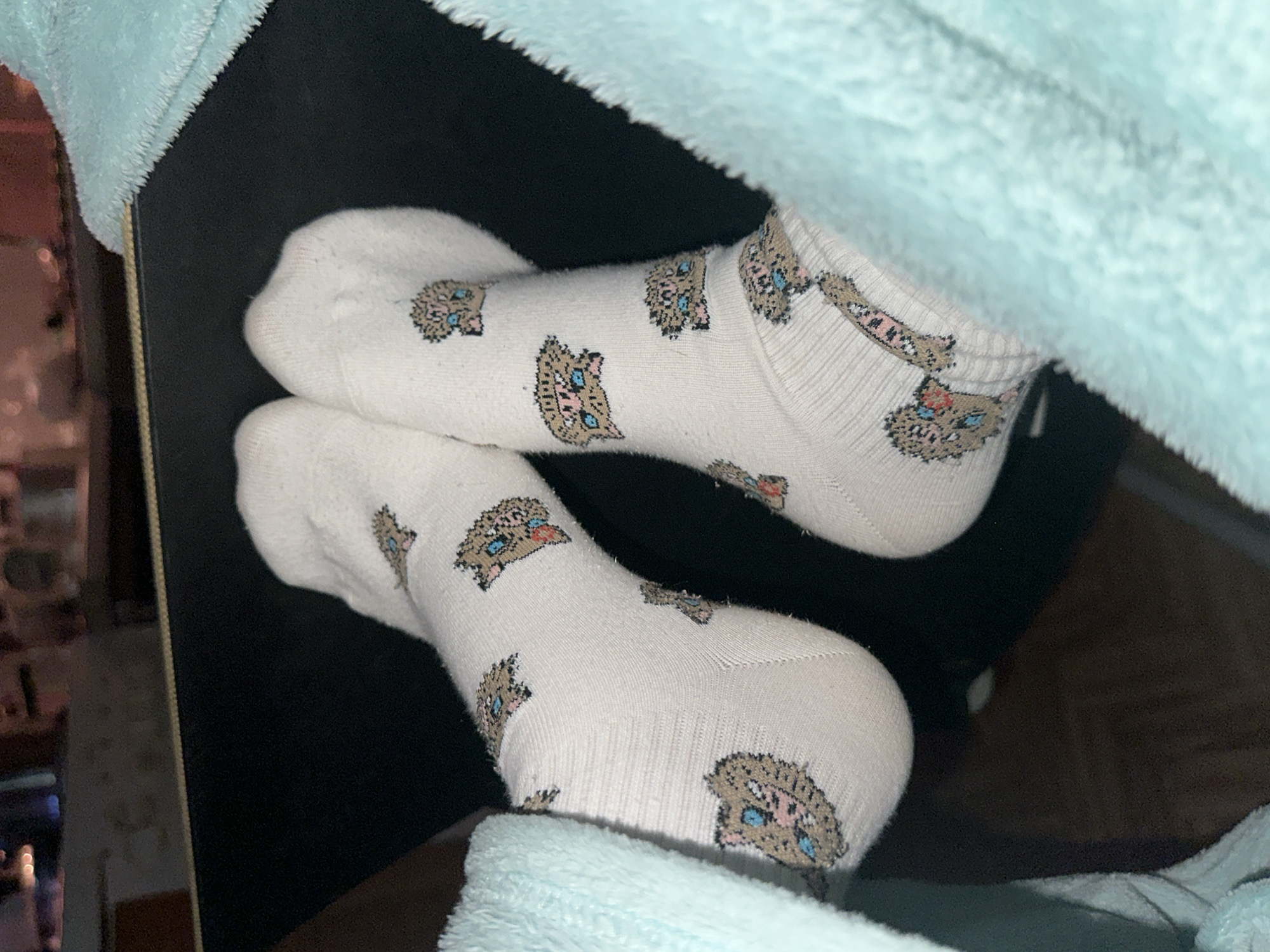 The scene is an indoor environment, located in Moscow, Russia. A pair of human feet, clad in white socks adorned with a repeating pattern of cartoonish brown, pink, and light blue boar-like heads, rests horizontally on a dark, flat surface. The feet are partially enveloped by a light blue, plush fabric, likely a blanket or robe, visible on both sides of the image. The dark resting surface features a subtle golden-brown trim along its visible upper edge. The background is out of focus, dimly lit, and shows indistinct shapes characteristic of an indoor setting, without specific identifying features.