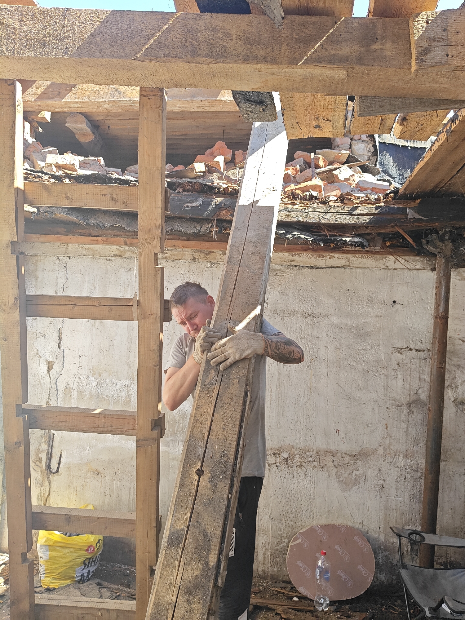 A man wearing gloves and a gray t-shirt holds a weathered wooden plank in front of a partially demolished building. The building appears to be in disrepair, with exposed brick, wood beams, and debris scattered around. A wooden frame or ladder-like structure stands beside the man. A yellow bag is visible near the base of the structure. Other objects, including a circular piece of cardboard, a water bottle, and a folding chair are in the scene. The location may be Chaikovsky, Russia.