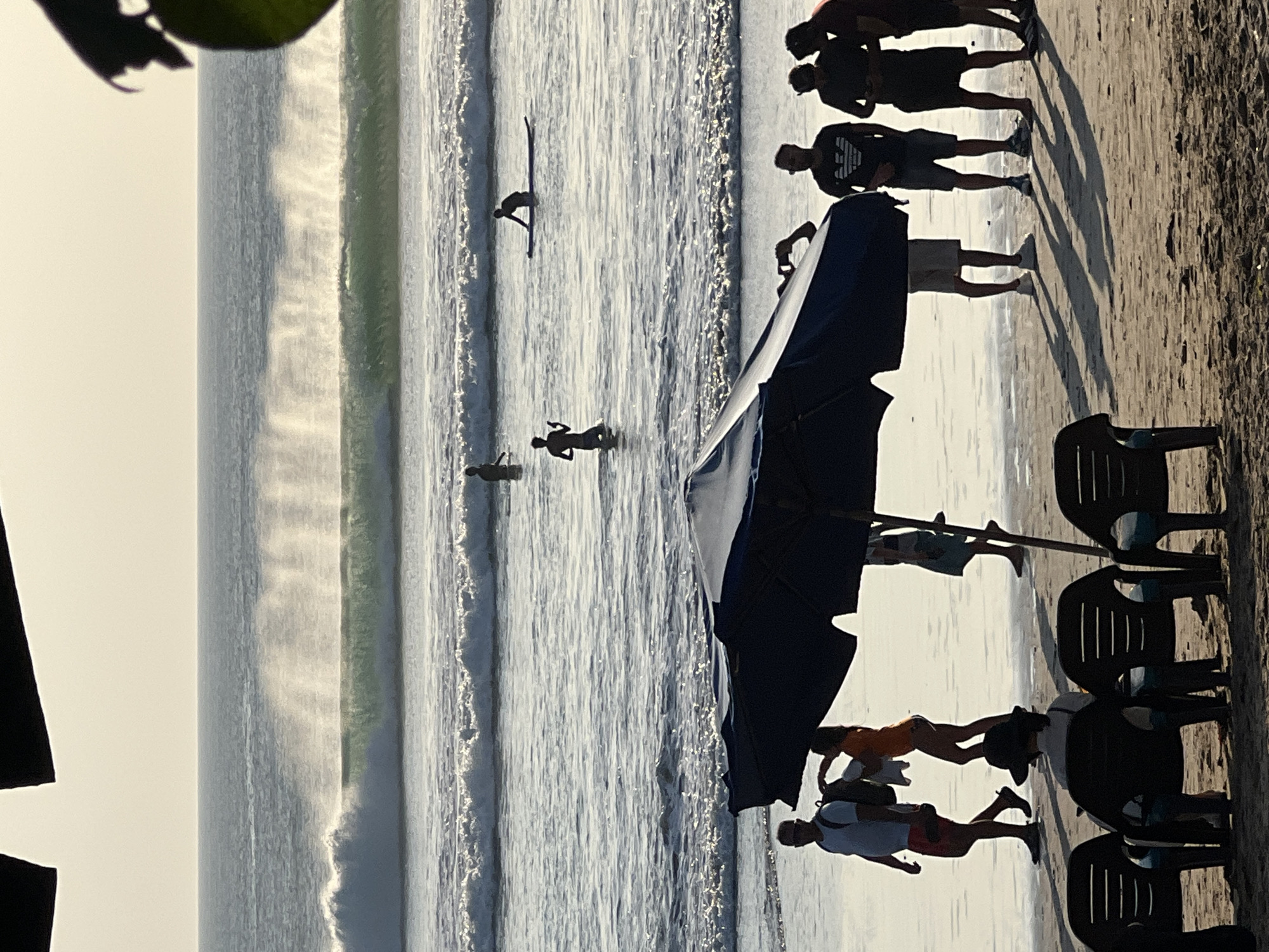 The image depicts a beach scene in Kuta, Indonesia, characterized by a wide, sandy shore and the ocean. The sun is low in the sky, casting long shadows and creating a bright, hazy atmosphere. In the foreground, several beach chairs are set up on the sand, some occupied and some empty.
A large, dark blue umbrella is positioned centrally in the mid-ground, providing shade. Several people are visible on the beach and in the water. On the shore, a man in a white t-shirt and red shorts walks towards the left, carrying a backpack. A woman in an orange top and bikini bottoms is next to him, adjusting a white towel. Further to the right, a group of men stand near the umbrella, some engaged in conversation.
In the water, several individuals are present. One person is wading in the shallows, while another is on a surfboard further out. A larger wave is breaking in the background, with its crest dissipating into a spray of mist. The ocean extends to the horizon, meeting a pale sky. The overall impression is of a relaxed, late afternoon at a popular beach destination.