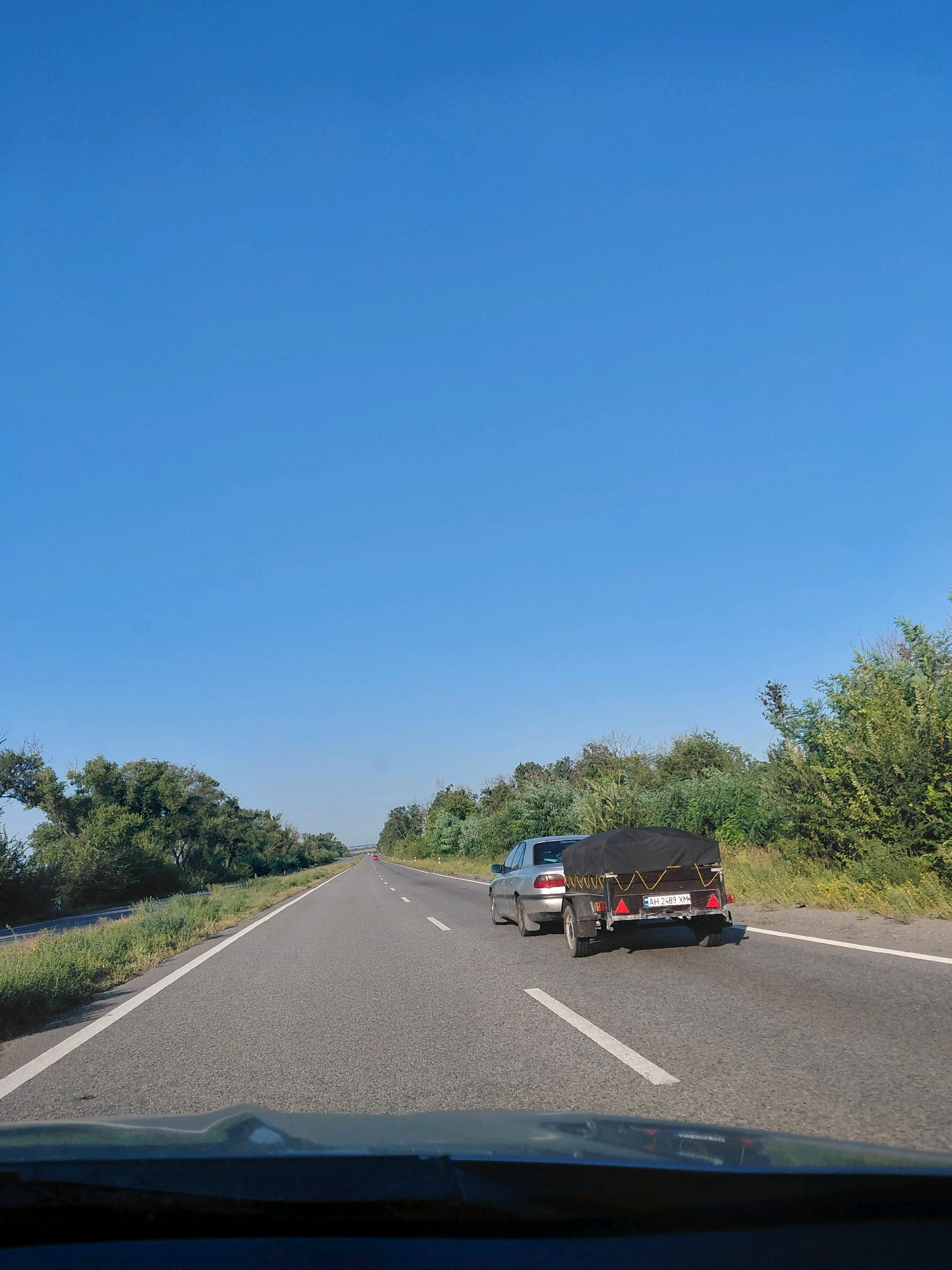 A silver sedan towing a black-covered trailer drives on a two-lane asphalt road through a rural landscape under a clear blue sky. The car and trailer are in the right lane, moving away from the viewer. The road is bordered by green vegetation and trees on both sides. The sun is bright, casting light on the scene. The image appears to have been taken from another vehicle traveling in the same direction.