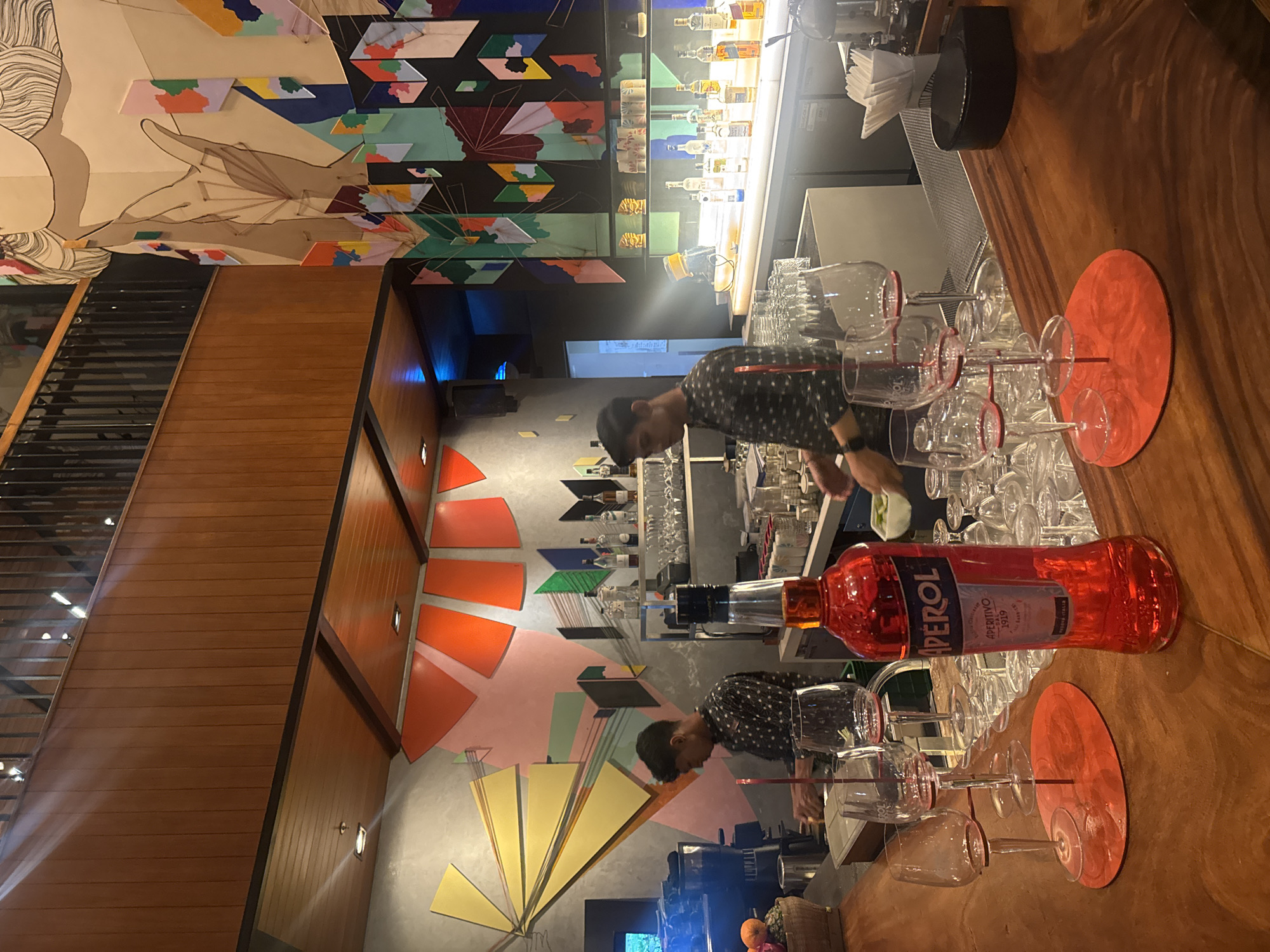 The image depicts the interior of a bar in Kuta, Indonesia. In the foreground, a wooden bar top displays a bottle of Aperol, several wine glasses on a red holder, and other glassware. Two bartenders are visible behind the bar. One bartender, wearing a black patterned shirt, is facing left and appears to be preparing drinks. The other bartender, partially obscured, is also behind the bar. The bar area is decorated with colorful abstract art on the walls. Behind the bar, shelves are stocked with various bottles of alcohol and more glassware. The lighting suggests an evening ambiance.