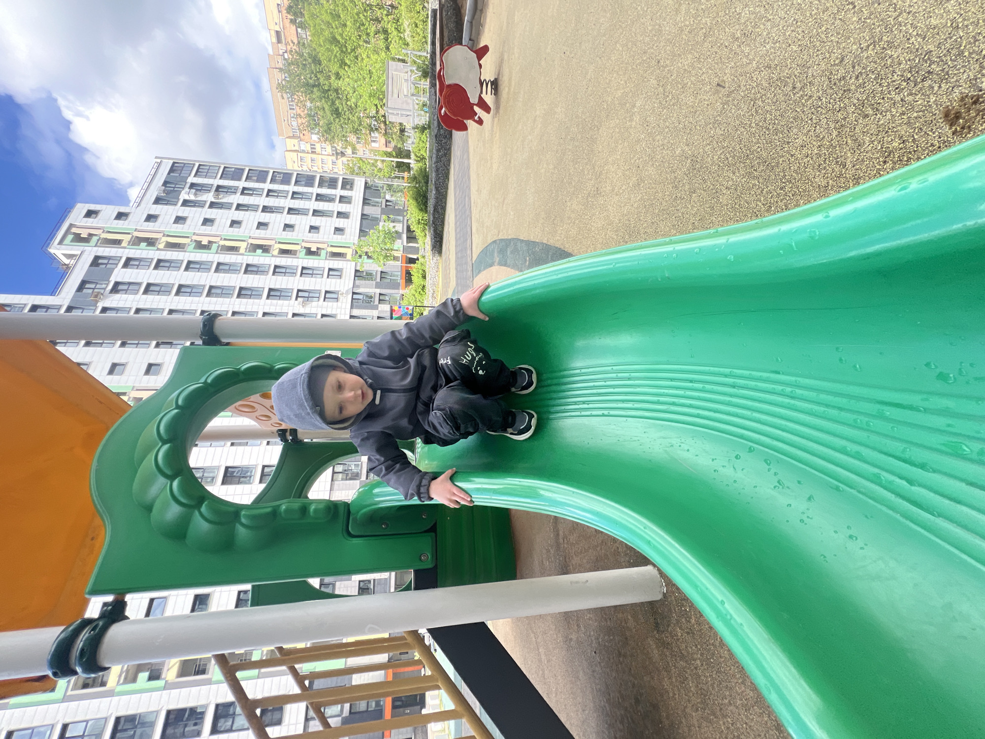 A young child is sitting at the top of a green slide in a playground. The child is wearing a dark hooded jacket and dark pants with white designs. Behind the slide, there's playground flooring and in the background, buildings can be seen, including a tall, modern apartment building with many windows. In the distance, another playground item, a red and white spring rider, is visible. The image suggests an outdoor playground setting. The location is Mytishchi, Russia.