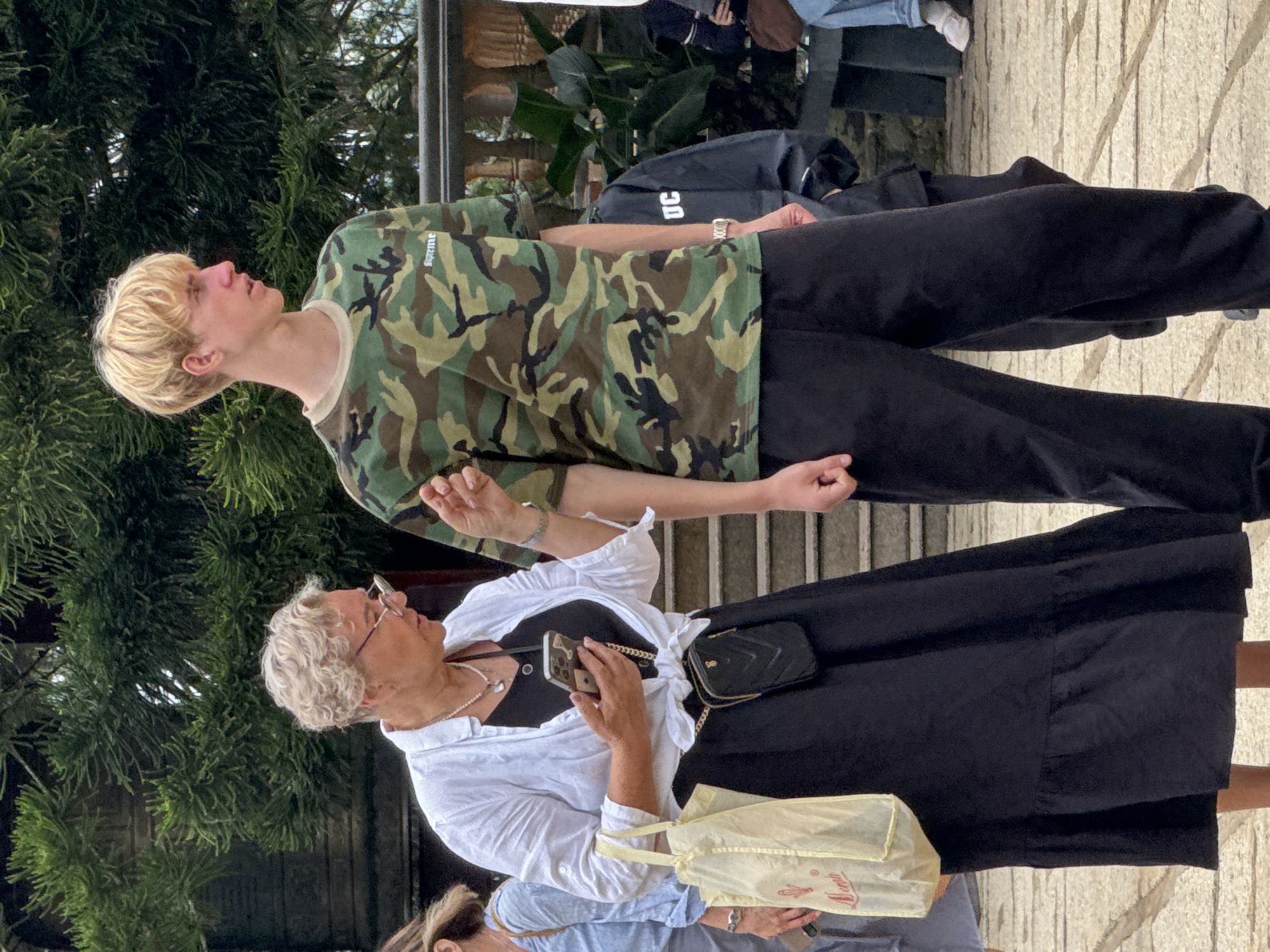 In Da Lat, Vietnam, on a paved walkway with stone stairs and lush greenery in the background, a Caucasian woman with blonde curly hair and glasses, wearing a white unbuttoned shirt over a black dress and holding a phone, stands to the left of a tall Caucasian male with short blonde hair, who is wearing a camouflage t-shirt and black pants. The woman is looking at her phone, and her hand is extended towards the man. A light yellow tote bag is hanging from her left arm. Behind the main subjects, another person in a dark jacket with "UC" visible on it is partially obscured. The overall scene captures a moment of casual interaction in an outdoor setting.