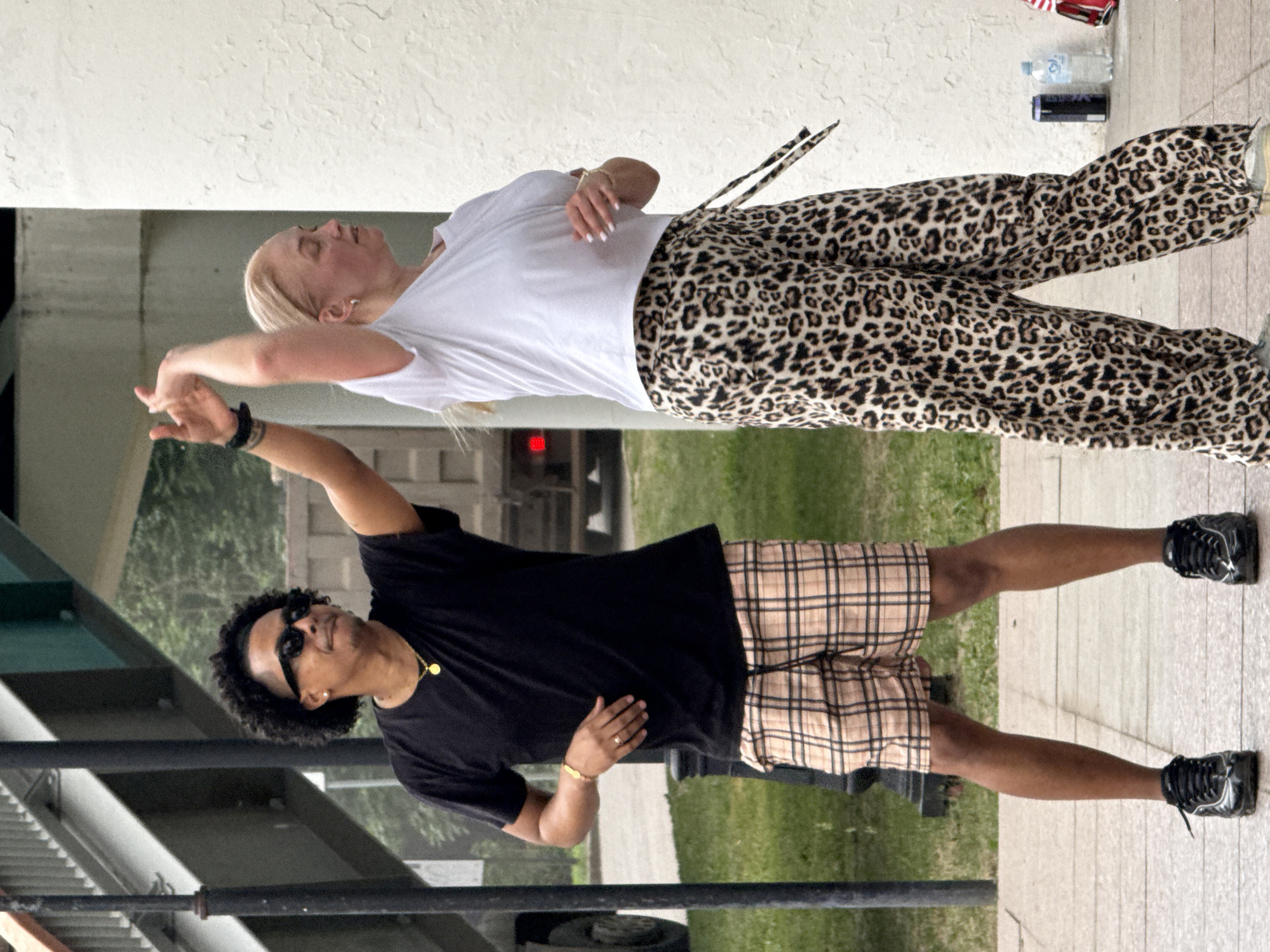 A man and a woman are dancing outdoors. The man, who appears to be of African descent, wears a black t-shirt, plaid shorts, and black sneakers. He has sunglasses on and is wearing a gold chain and a bracelet. The woman, who is blonde, wears a white t-shirt and leopard-print pants. She has on hoop earrings and bracelets. They are positioned on a paved area in front of a white textured wall and a grassy area. In the background, there is a bridge, a pole, and a truck. There are also some bottles and a bag on the ground. The scene appears to be in an urban environment, possibly a park or public space.