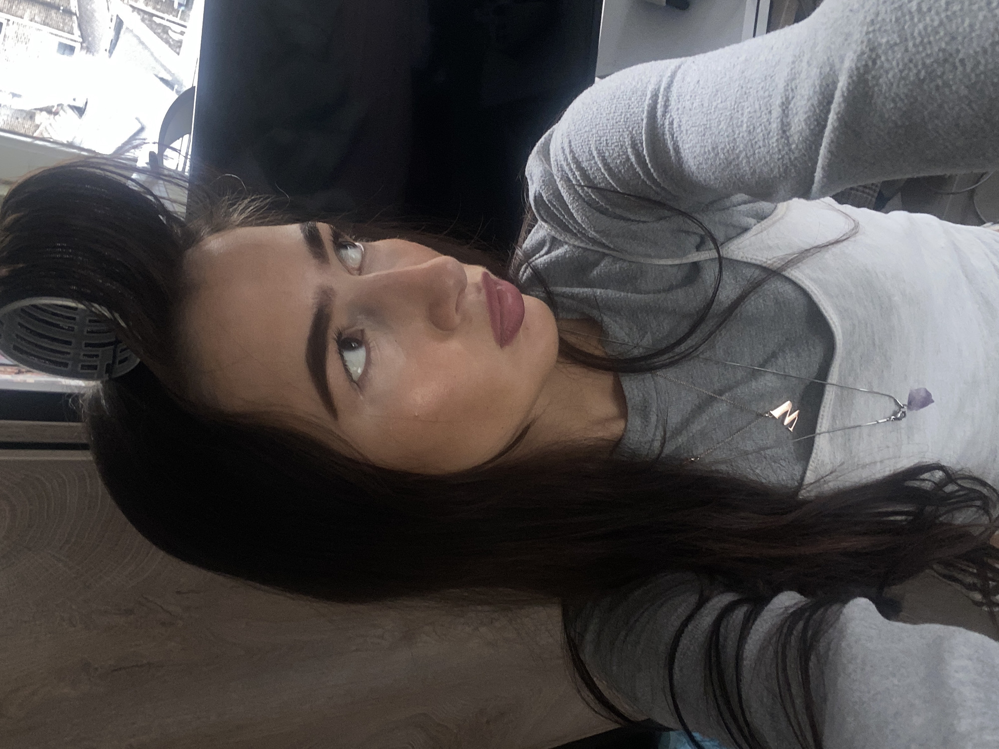 The image is a selfie taken at an angle, featuring a woman with long, dark brown hair. Part of a gray plastic hair roller is visible in her hair. She is wearing a gray sweatshirt over a white top, and a silver necklace with a letter "M" pendant and a purple stone is around her neck. The background shows part of a television screen and a blurred view of a city through a window, possibly the city of London based on building styles and sky conditions. The photo is taken indoors, possibly in a home setting, with a light-colored wooden surface visible at the bottom of the frame.