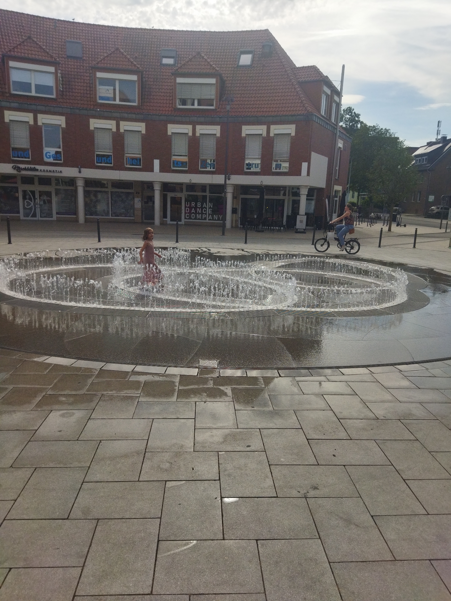 This is a full-shot, eye-level, outdoor photograph of a public square in Monheim am Rhein, Germany, featuring a circular fountain with multiple jets spraying water. A young girl in a light-colored dress is standing in the fountain, her body partially obscured by the water. To the right of the fountain, a woman is riding a bicycle. In the background, there is a brick building with multiple stories and windows, and a sign that reads "URBAN DANCE COMPANY". The ground is paved with large, rectangular stone tiles. The sky is overcast with some patches of blue.