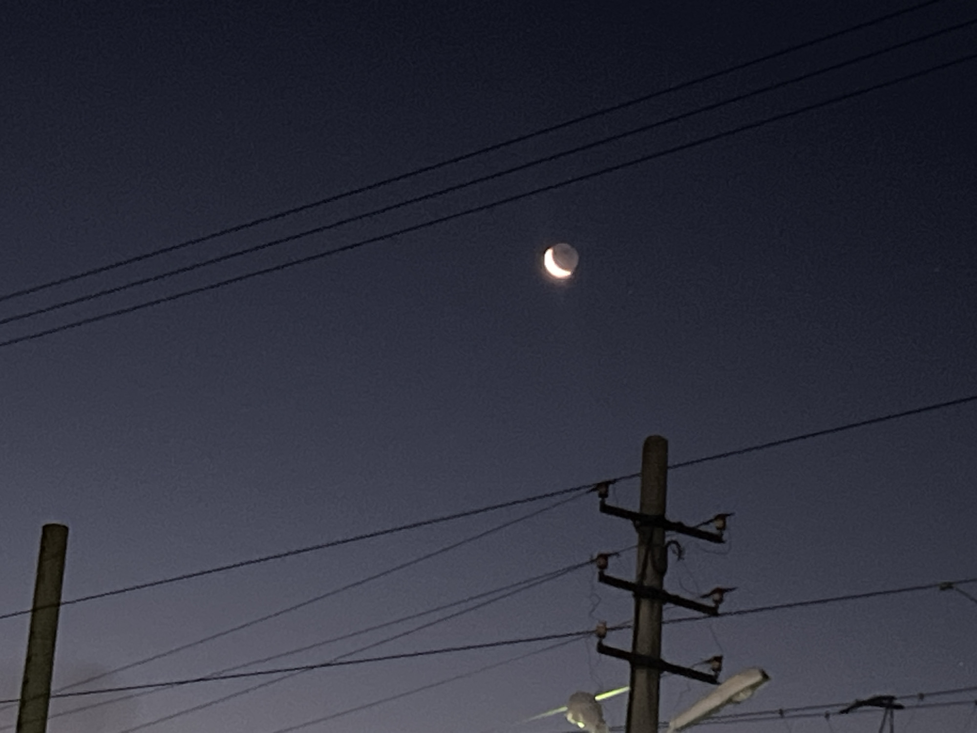 A crescent moon is visible in the dark sky, positioned above three horizontal power lines. In the foreground, a wooden utility pole with insulators and wires is present. Another vertical pole is visible on the left side of the image. The scene appears to be an outdoor urban environment at dusk or night, possibly Iksha, Russia.