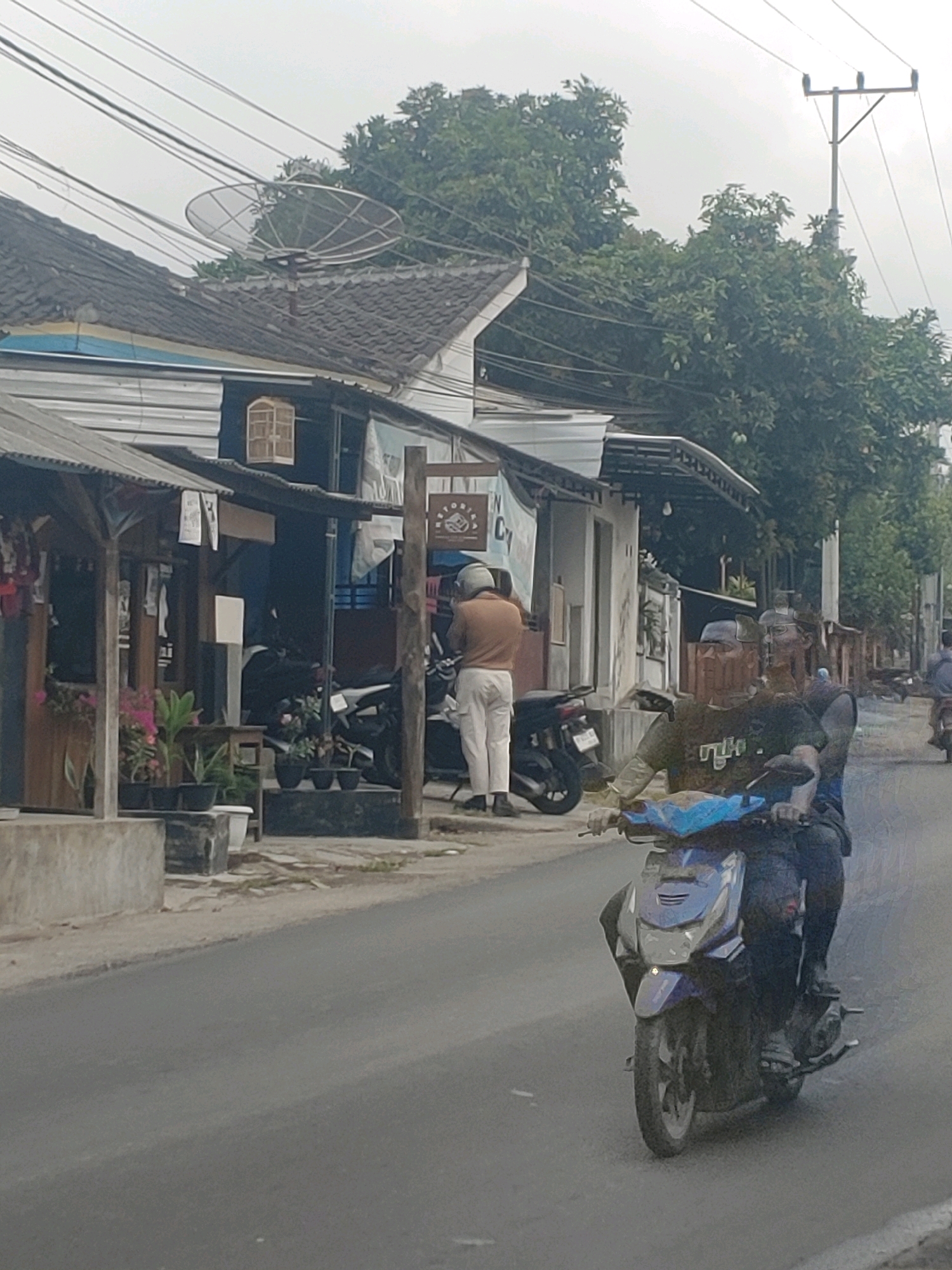 The image depicts a street scene in Kembang Kerang, Indonesia. In the foreground, two men are riding on a blue and silver motorcycle. The rider is wearing a black t-shirt with white lettering and dark pants, while the passenger behind him is wearing a dark t-shirt and dark pants. Both appear to be wearing head coverings. The motorcycle is moving on an asphalt road.
In the middle ground, there are several buildings with awnings and open fronts, suggesting a commercial area. One building features a satellite dish on its roof. A person in a light-colored shirt and light-colored pants is standing next to a motorcycle parked in front of one of the shops. Various potted plants are visible near the buildings.
The background shows trees and a power pole with electrical wires. The sky is overcast. The overall impression is a glimpse into daily life on a street in a town or city in Indonesia.