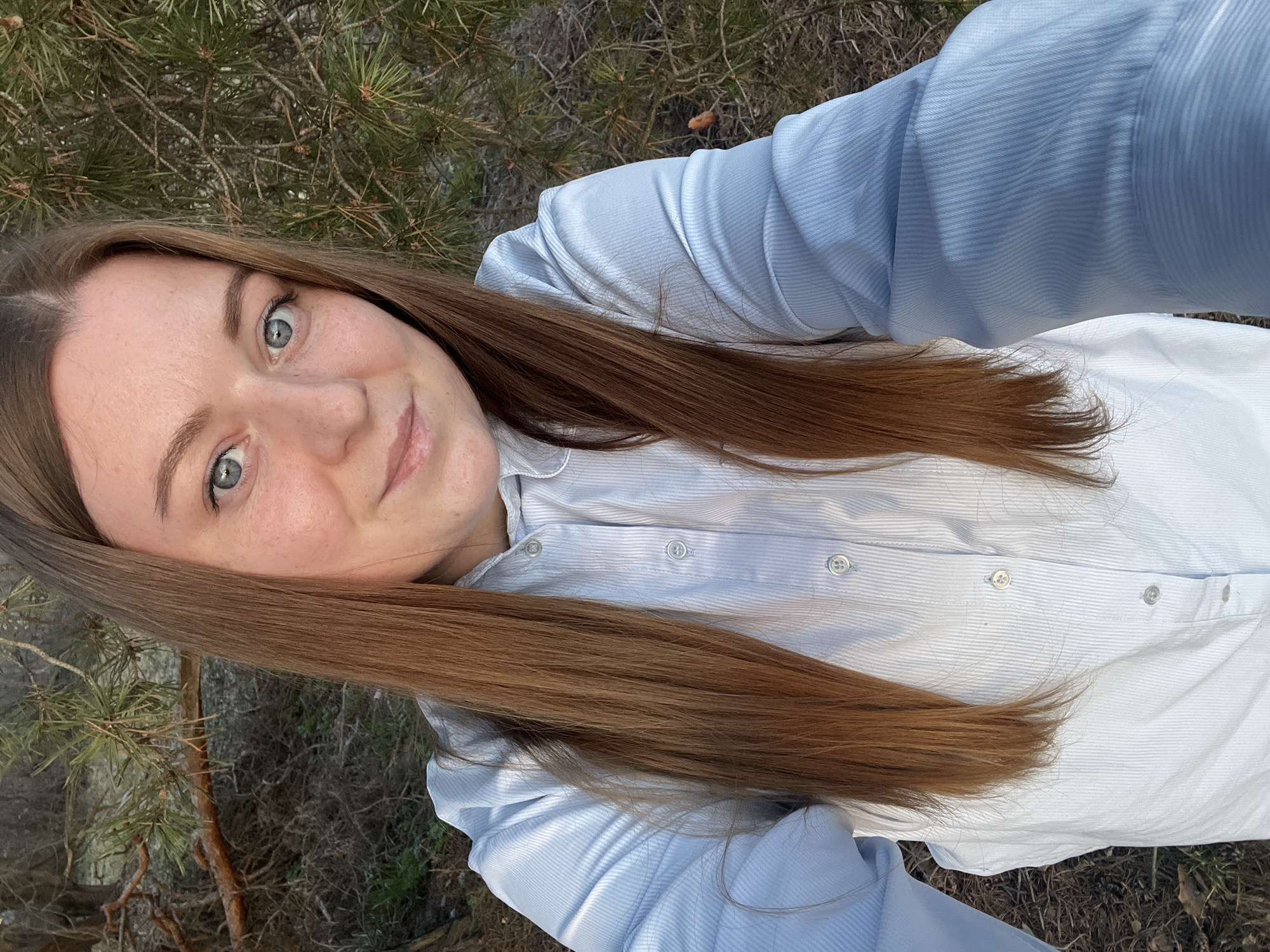A woman with long, auburn hair and light blue eyes is positioned in front of an outdoor scene, likely in a natural setting. She is wearing a light blue, long-sleeved, button-down shirt. The background includes dense foliage and possibly pine trees, suggesting a forested or natural area. The image is taken from a slightly angled perspective, as if the woman is holding the camera herself for a selfie. The location is indicated to be in Раздолье, Russia.