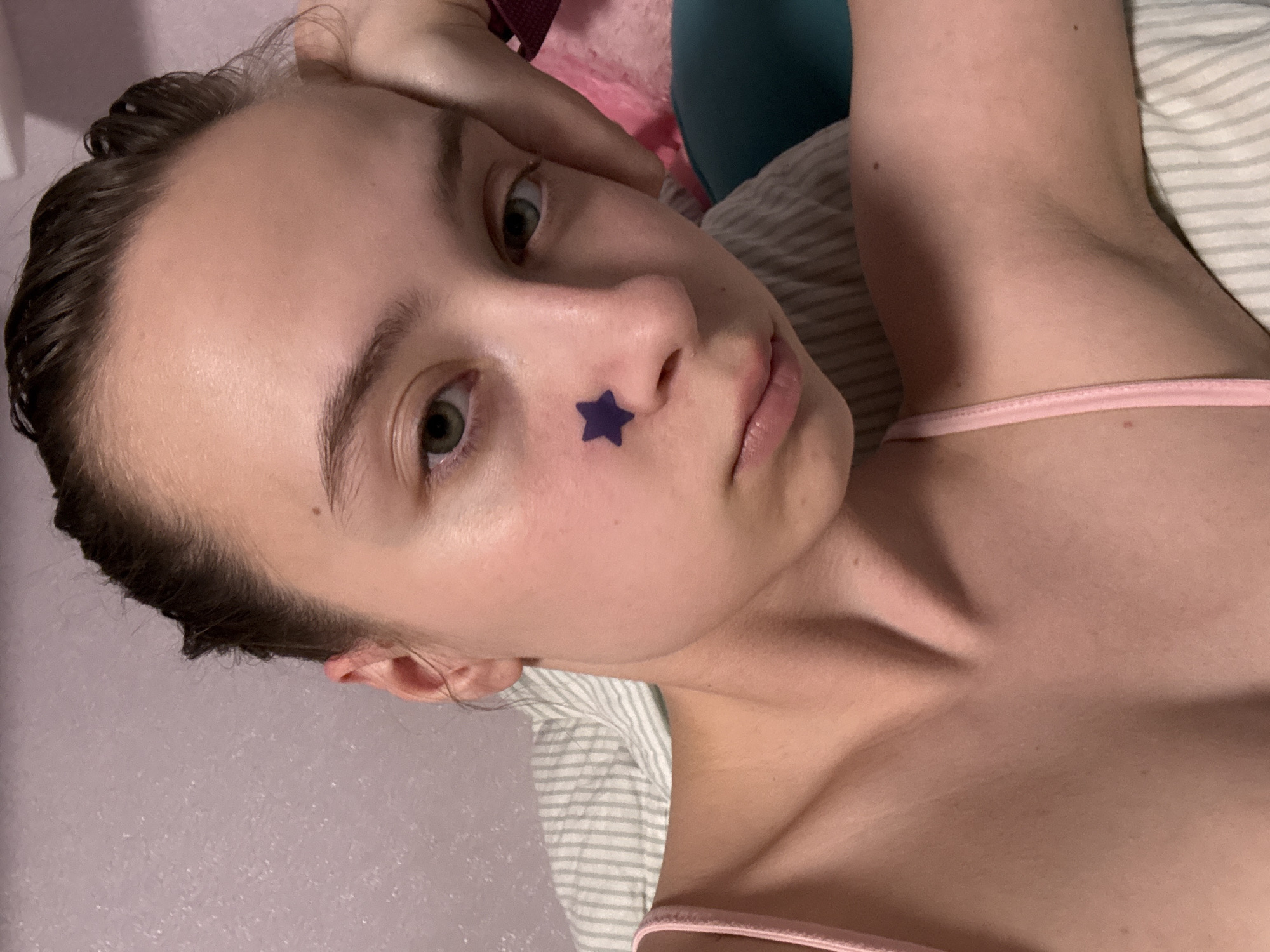 A young woman is lying down. She has short brown hair, fair skin, and green eyes. A dark purple star-shaped sticker is on her nose. She is wearing a pink spaghetti strap top and is lying on a white and grey striped surface that appears to be bedding. A pink and teal object is behind her head. The background is a light pink wall.
There is no information to place the scene in Прикамский, Russia.