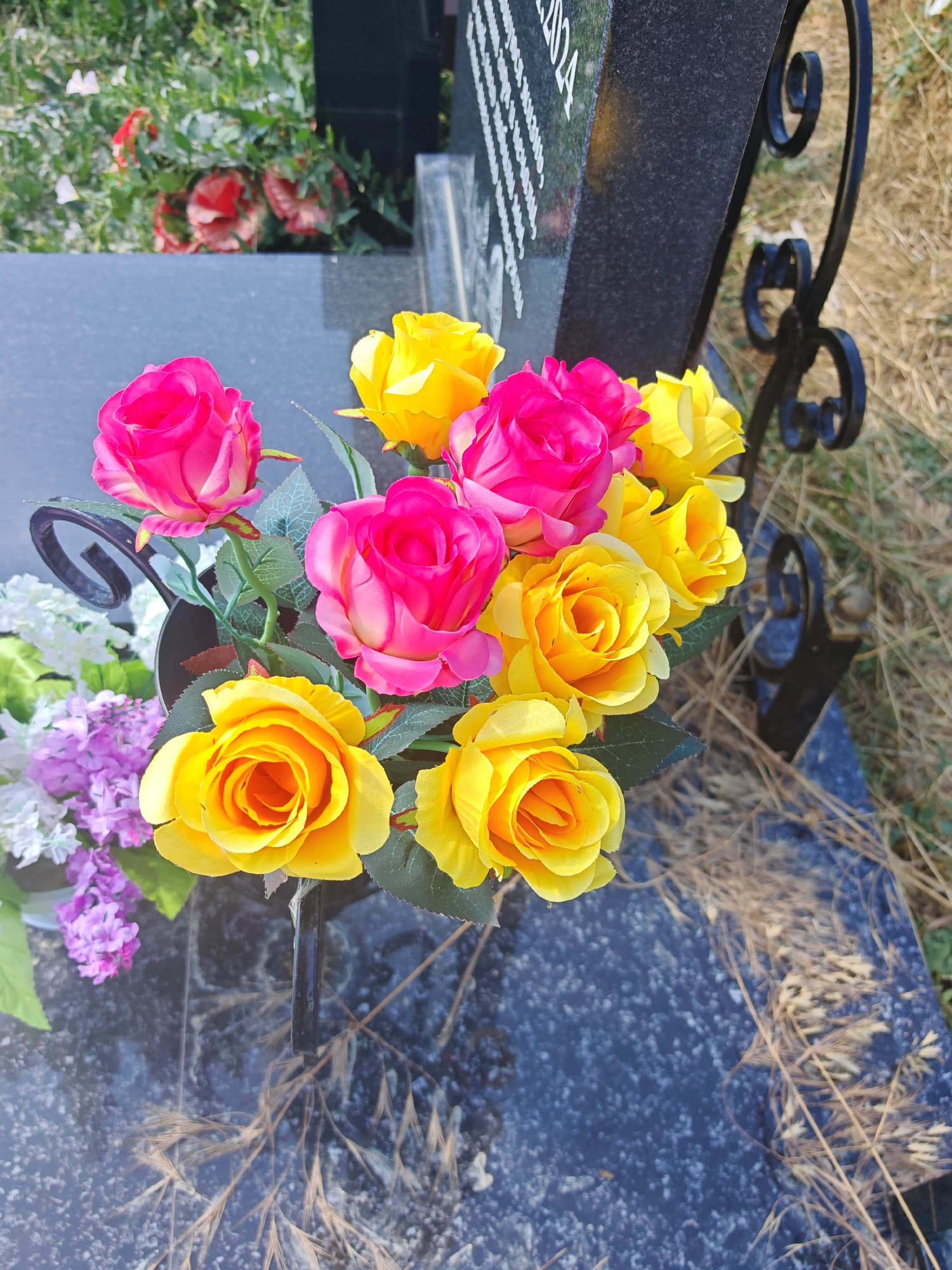 The image shows a close-up view of a bouquet of artificial roses, predominantly yellow and pink, placed in a black metal holder on a grave. The flowers are arranged in front of a dark gray granite headstone, part of which is visible with some text etched into it. The headstone is positioned on the ground, which appears to be covered with dry grass and some greenery. A decorative black metal fence with scrollwork is also visible to the right of the headstone, partially obstructing the view of the grave surroundings. The lighting suggests it is daytime, with natural sunlight casting shadows and highlighting the textures of the flowers and the stone. The image appears to be taken in a cemetery. Based on the information, this scene is located in the city of Supsekh, Russia.