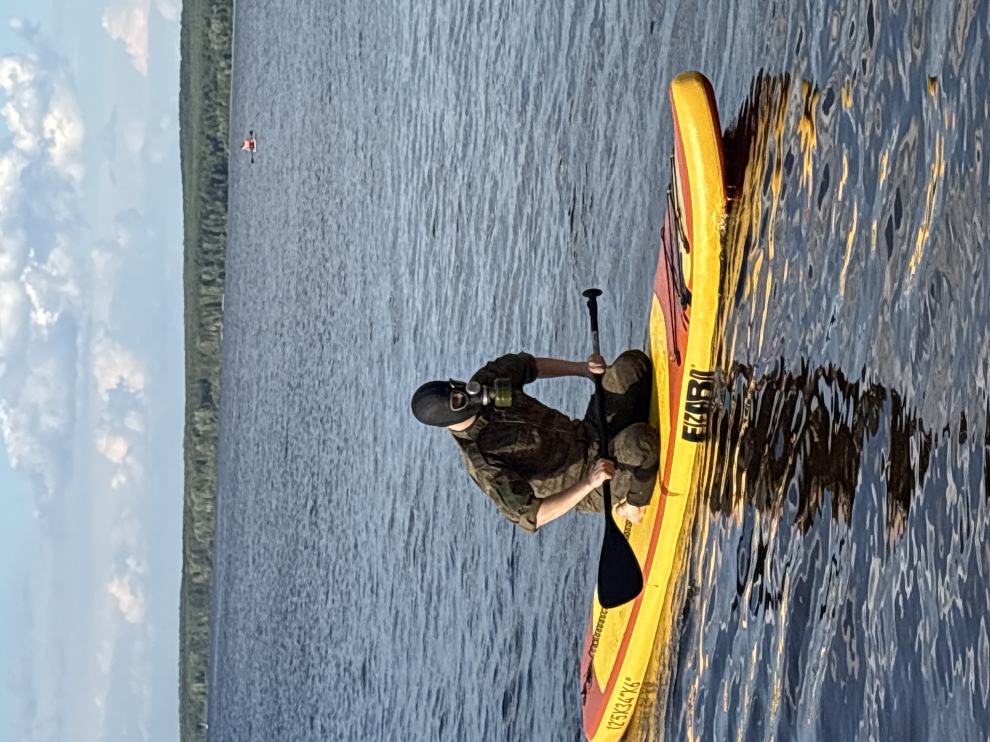 A person wearing a black balaclava and a green gas mask is kneeling on a yellow and red paddleboard in a body of water, holding a paddle. The person is wearing a camouflage-patterned shirt and pants. The paddleboard has the brand name "FIZARI" visible. The background shows a calm blue lake with a dense green forest on the horizon under a partly cloudy sky. The water is reflecting the sky and the paddleboard. The location is the Kama River in the city of Chaikovsky, Russia.