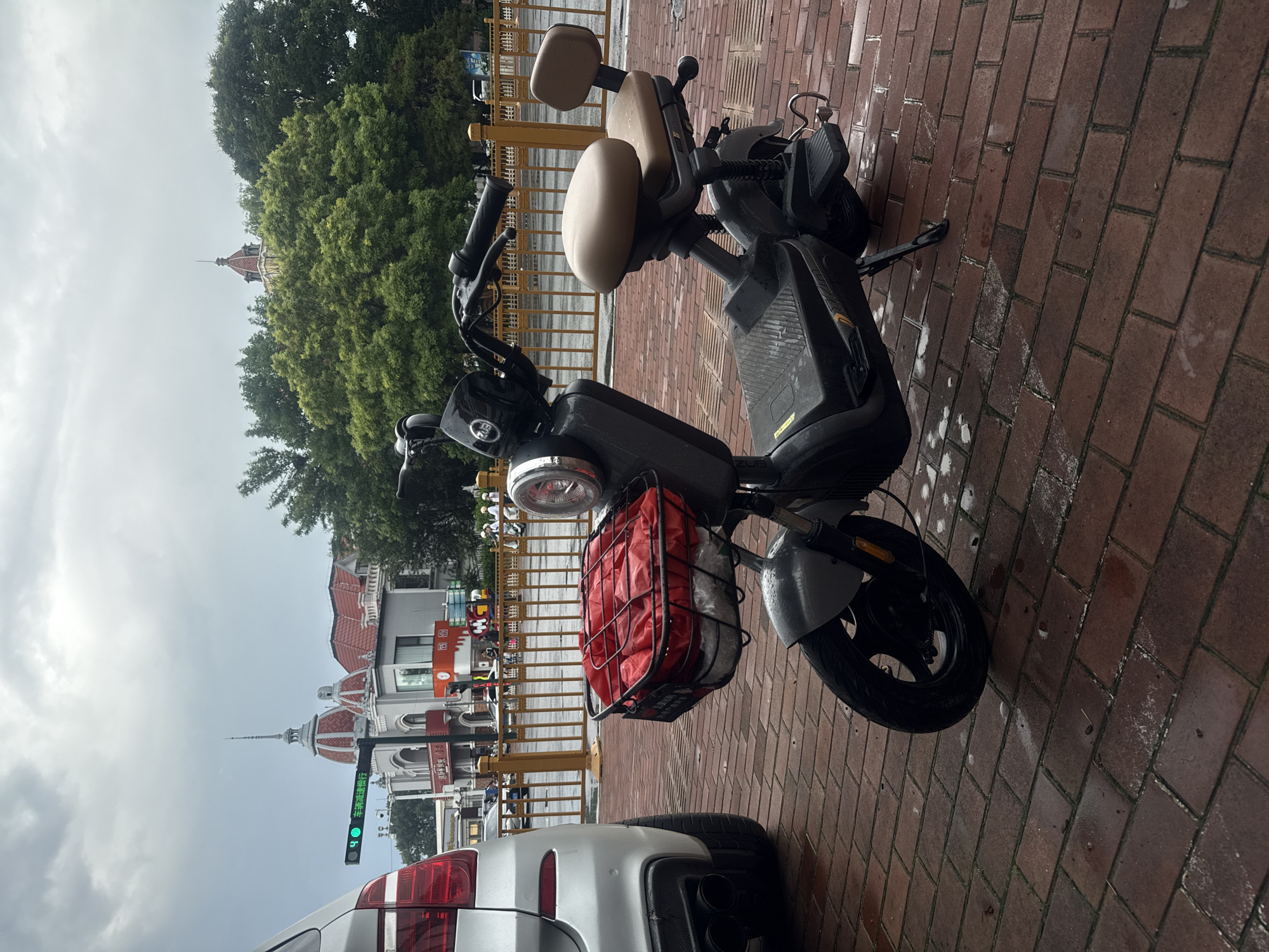 An electric scooter with a red basket filled with items and a brown seat is parked on a red brick sidewalk. To the left of the scooter is the rear of a silver car. In the background, there is a yellow metal fence, trees, and buildings with distinctive domed roofs. A traffic light with a green signal is visible on the left side of the image. The scene appears to be in Qinhuangdao, China. The sky is overcast.
