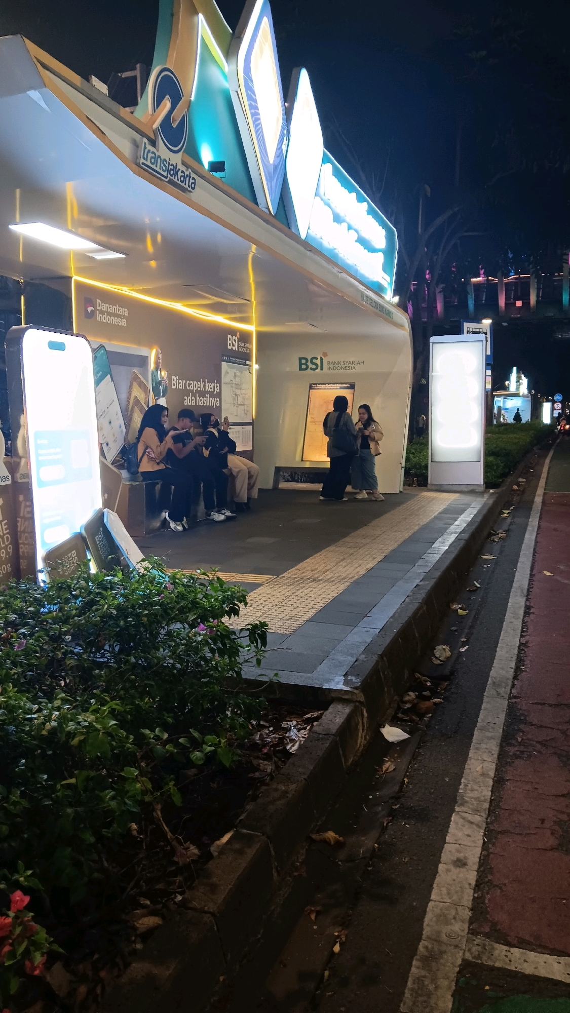 The image depicts a bus stop in Central Jakarta, Indonesia, at night. The bus stop features a modern, illuminated design with a TransJakarta logo prominently displayed. Several individuals are visible, with four people sitting on a bench, appearing to be looking at their phones. Two other individuals stand near a poster on the bus stop structure. The bus stop is situated next to a sidewalk and a street, with trees and other city lights visible in the background. The area appears to be clean, with some fallen leaves on the ground near the curb. The lighting suggests it is nighttime.