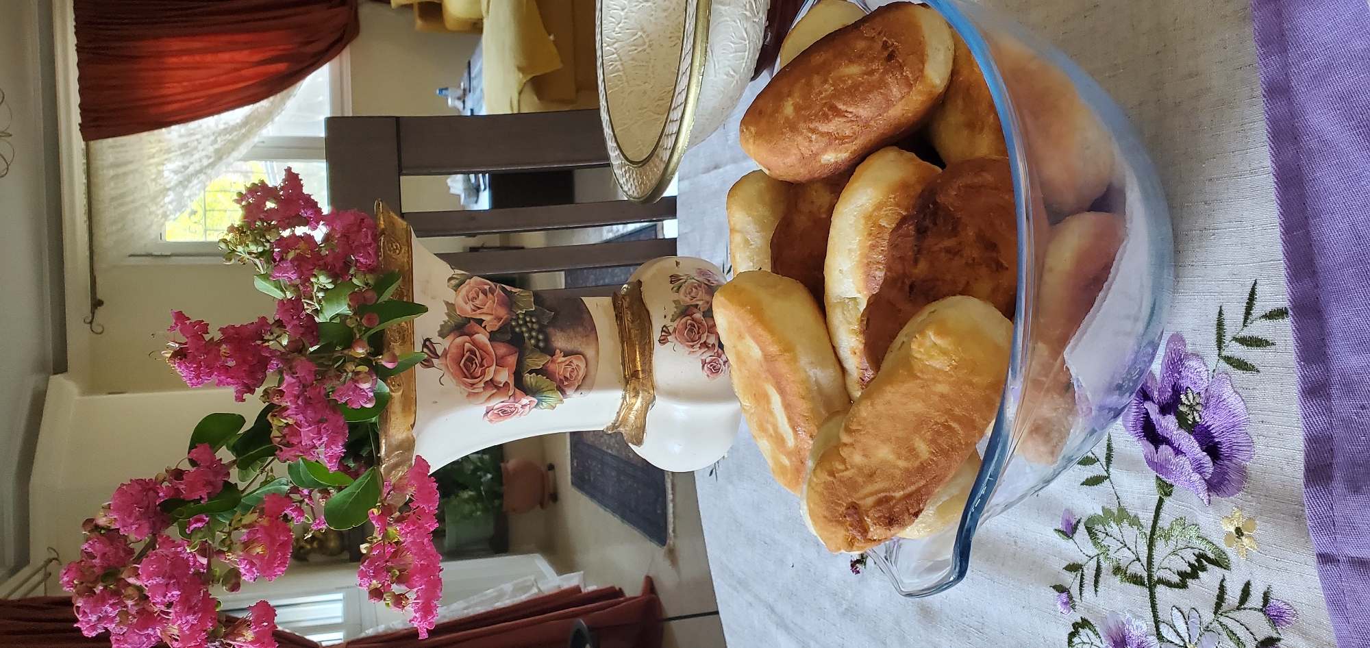 A close-up, eye-level shot captures a glass bowl filled with golden-brown, fried pastries, likely pirozhki or similar dough-based treats. The pastries vary in shape and color, some appearing lighter and fluffier, while others are more deeply browned. The bowl itself is clear glass with a rippled edge. The table is covered with a white tablecloth embroidered with purple floral patterns, and a darker purple fabric strip runs along the bottom edge of the frame.
Behind the bowl, a decorative ceramic vase with a gold rim and base is prominently displayed. The vase is adorned with hand-painted roses and grapes. It holds a vibrant bouquet of pink flowers with green leaves, adding a splash of color to the scene. The vase and flowers sit on the white tablecloth.
Further in the background, a dark wooden chair is visible, positioned at a table. Beyond the chair, a window with sheer white curtains and heavy, dark red drapes is present. The window offers a glimpse of an outdoor scene, possibly with greenery. A hint of a yellow sofa and a dark table can be seen in the distant background through the chair.
The overall setting suggests an indoor domestic environment, likely a dining or living area, with a focus on a freshly prepared food item and decorative elements. The lighting appears to be natural, coming from the window, creating soft shadows and highlighting the textures of the food and vase. The image does not contain any identifiable people or specific landmarks that would definitively place it in Νέα Σάντα, Greece, or any other specific city.